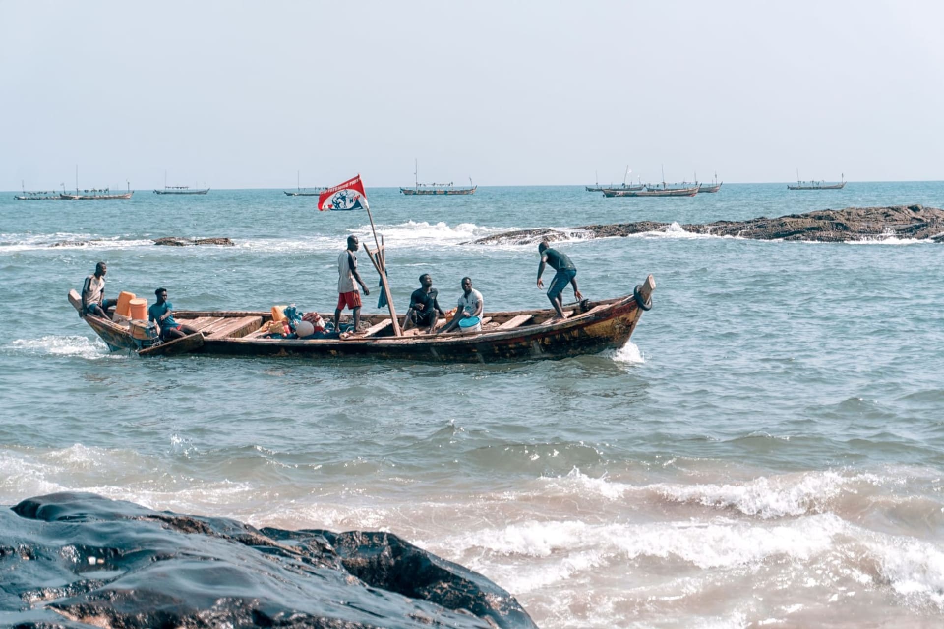 The whitewashed walls of Cape Coast Castle rising from a rocky promontory above the Atlantic, cannon ports facing the grey-green sea under a heavy coastal sky.