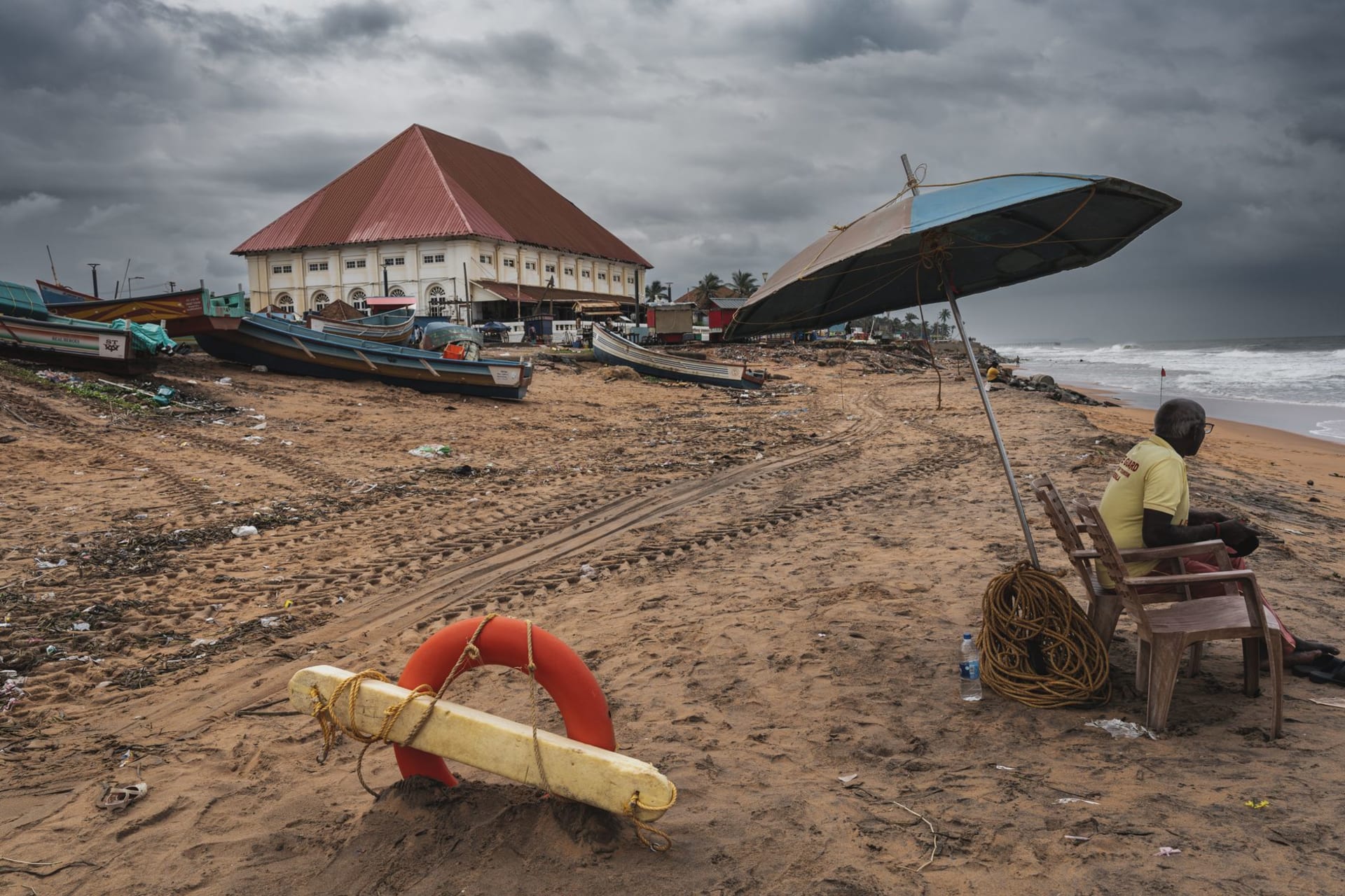 A lone surfer paddling out through green Atlantic waves at Busua beach, Ghana, with fishing boats visible on the sand behind them