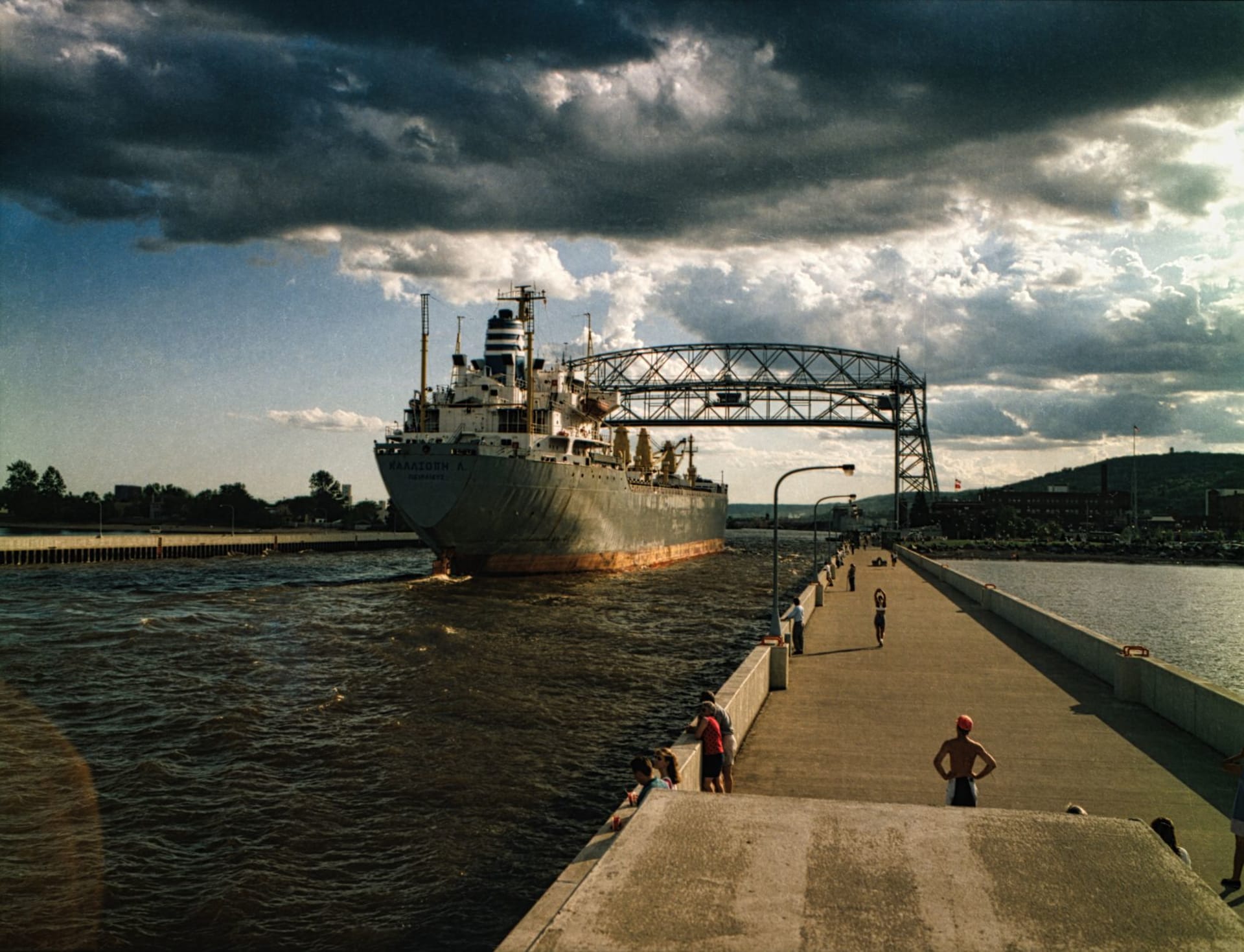 The Aerial Lift Bridge at Canal Park in Duluth with a massive iron ore freighter passing beneath it, crowd gathered along the promenade