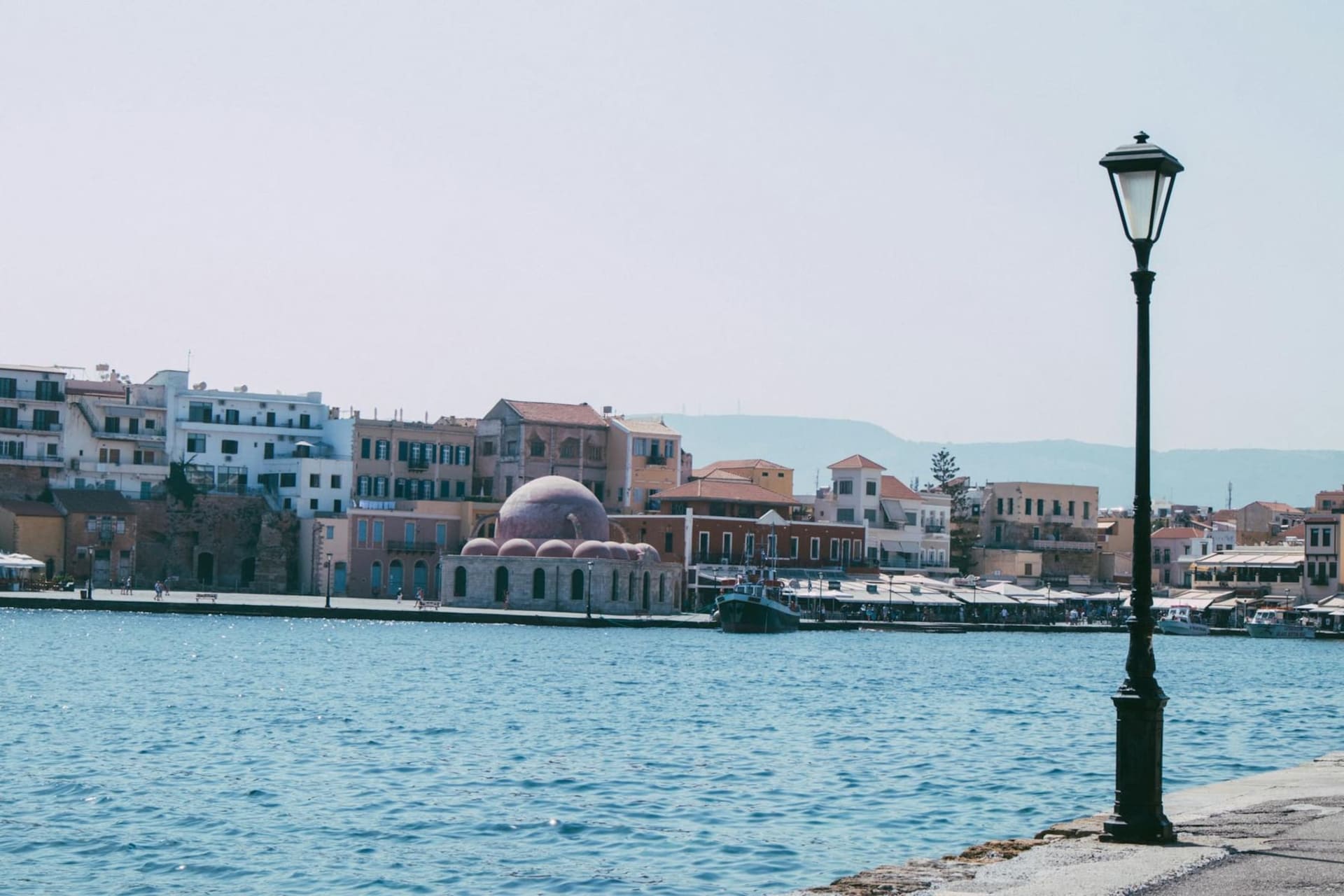The crescent Venetian harbor of Chania at dusk, pastel ochre and terracotta facades reflected in the still water, the domed Egyptian lighthouse anchoring the far end of the quay