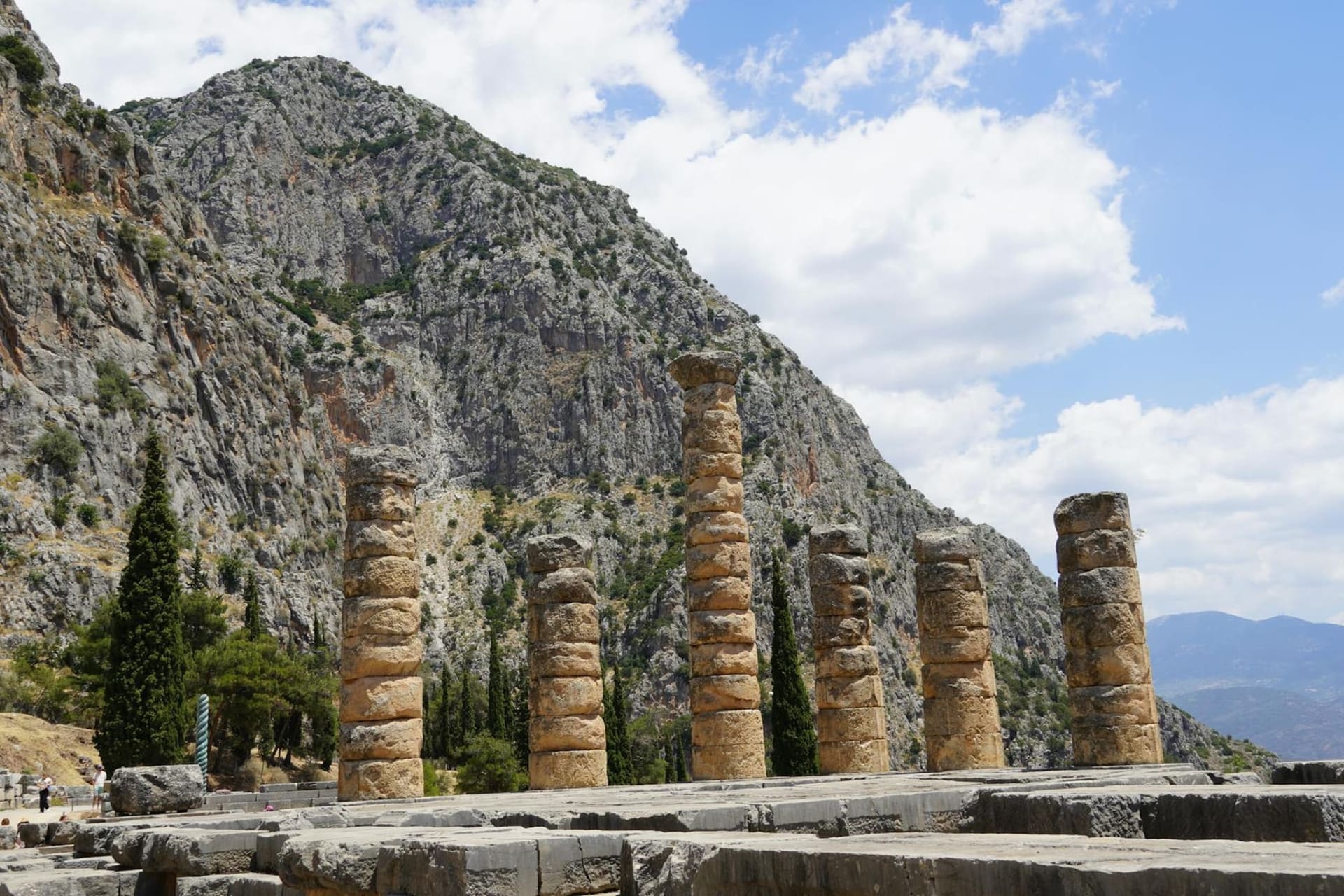 Ancient limestone columns of the Temple of Apollo at Delphi standing against a dramatic backdrop of steep olive-covered mountain slopes and a wide valley below