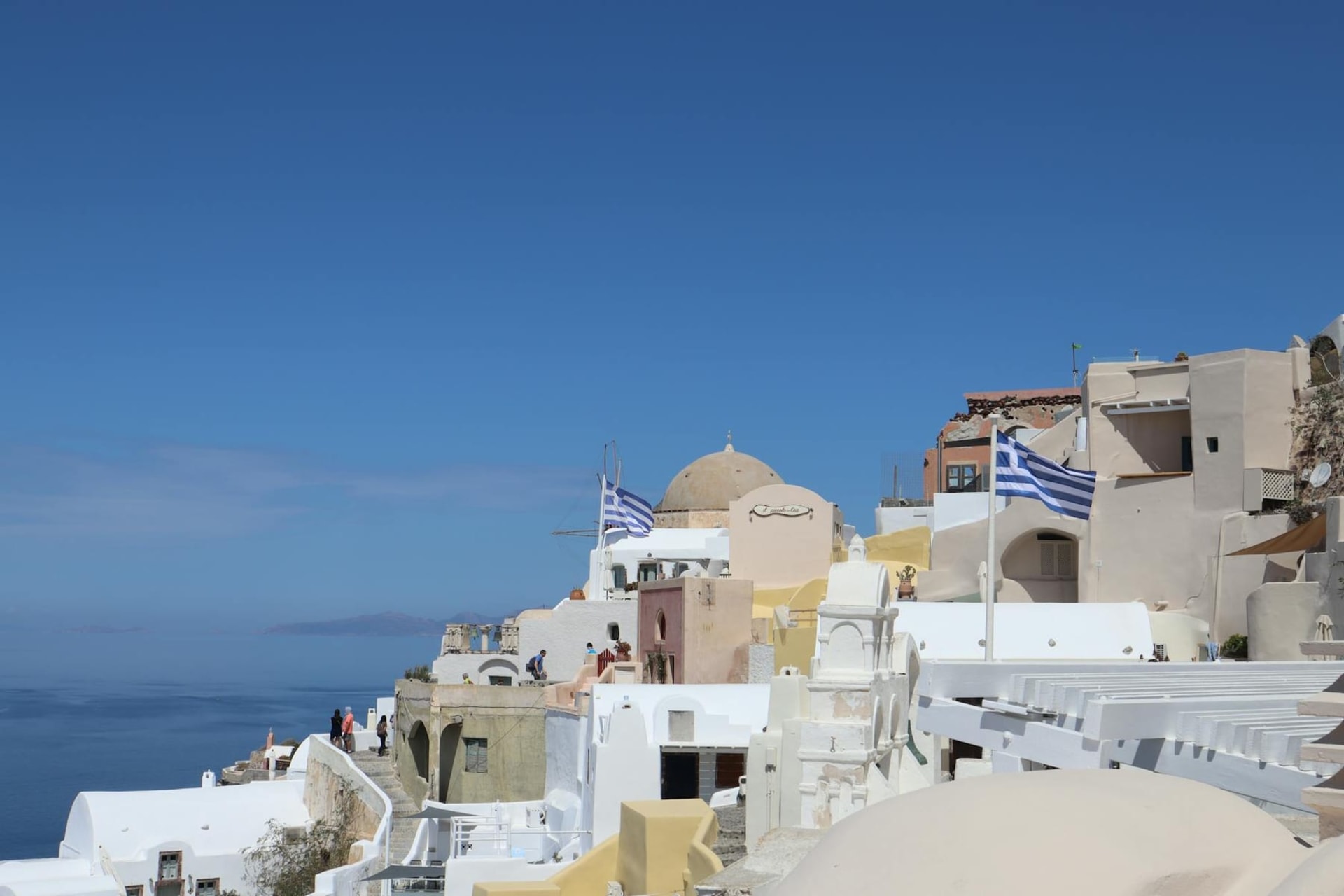 Whitewashed houses and a small blue-domed chapel perched on the volcanic cliffs of Thirasia, with the deep blue caldera and Santorini's distant skyline stretching behind them under soft Aegean light.