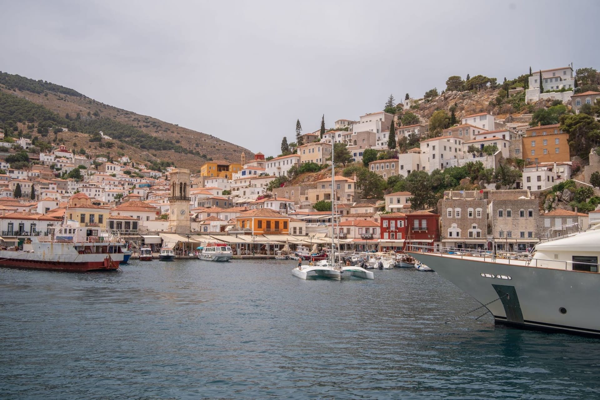 The stone harbor of Hydra Town at dusk, fishing boats moored along the quay and whitewashed captain's mansions climbing the hillside behind
