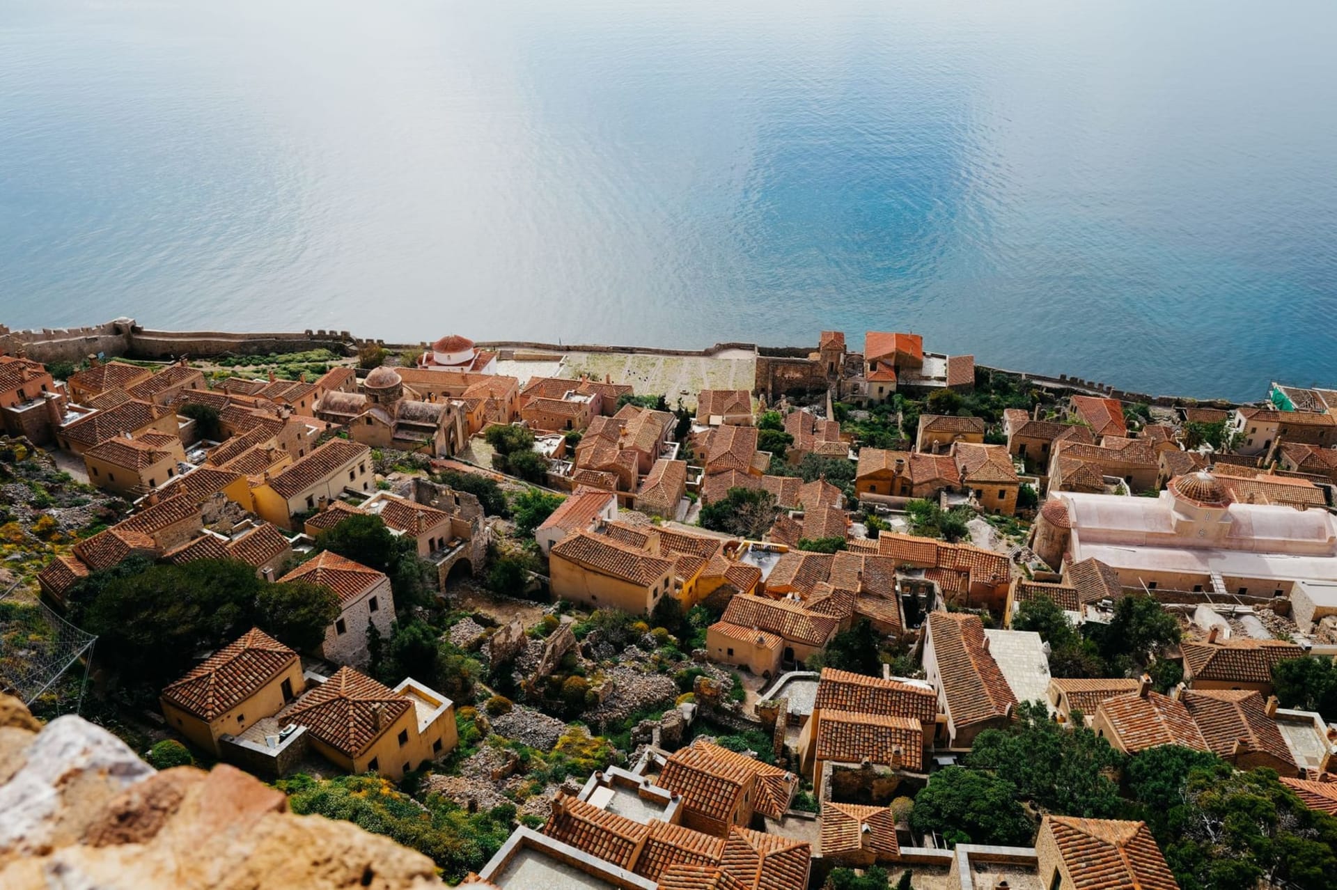 The great sea rock of Monemvasia rising from the Aegean at dusk, its Byzantine walls and red-roofed buildings stacked against the cliff face, reflected in still dark water below