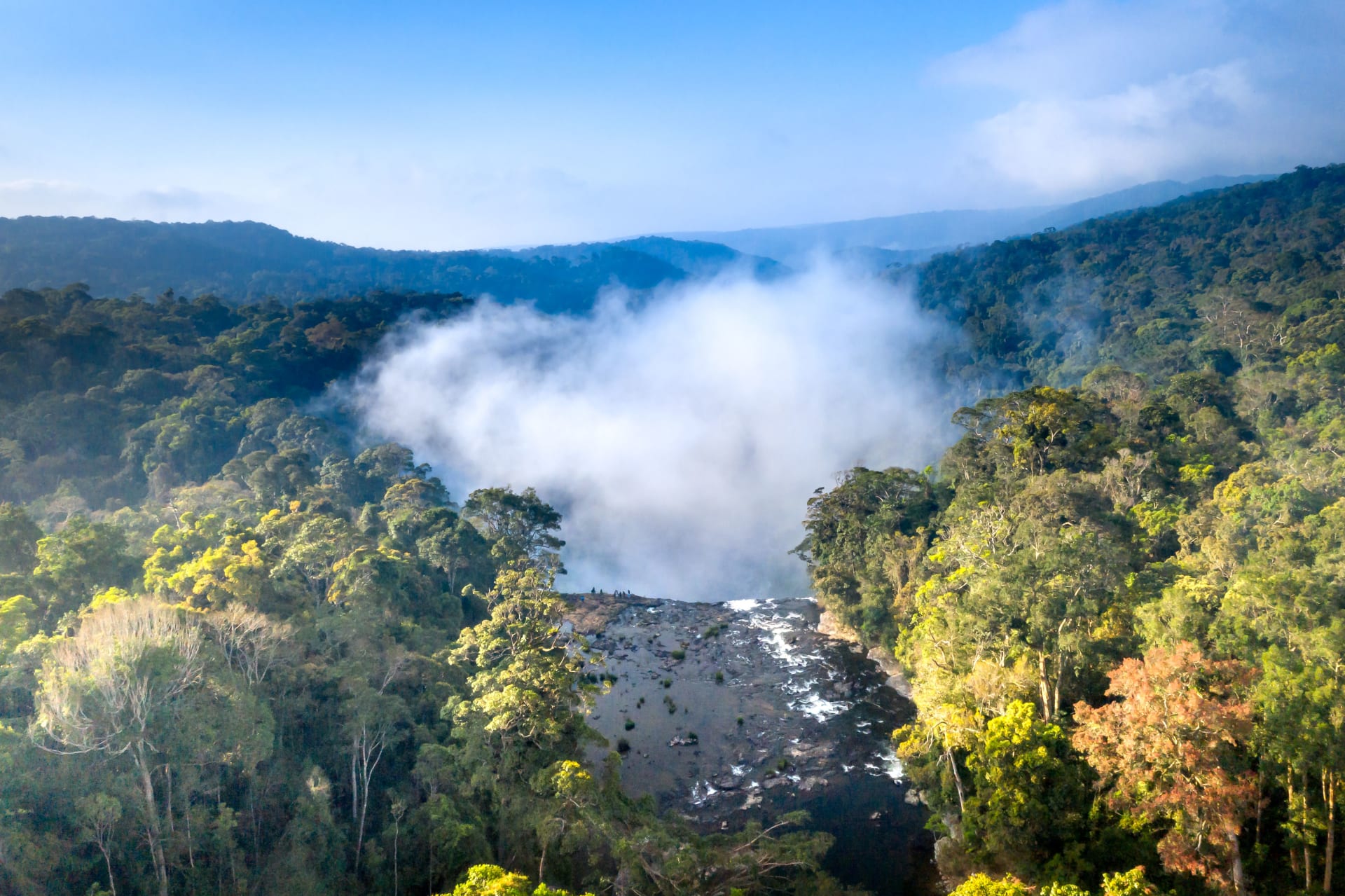Breathtaking aerial shot of a misty waterfall cascading into dense green jungle, clouds drifting through the forest canopy below