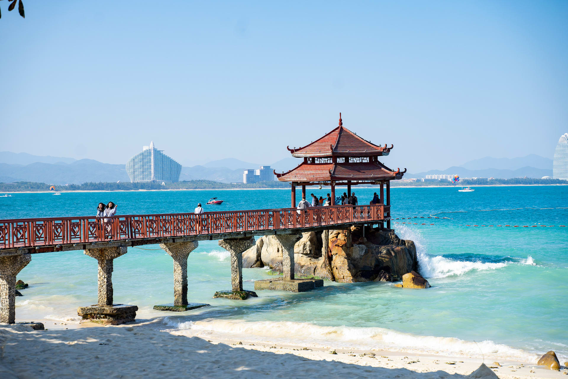 Haitang Bay beach at midday with clear pale-turquoise water, white sand, and the towers of the duty-free shopping complex visible behind a row of palms