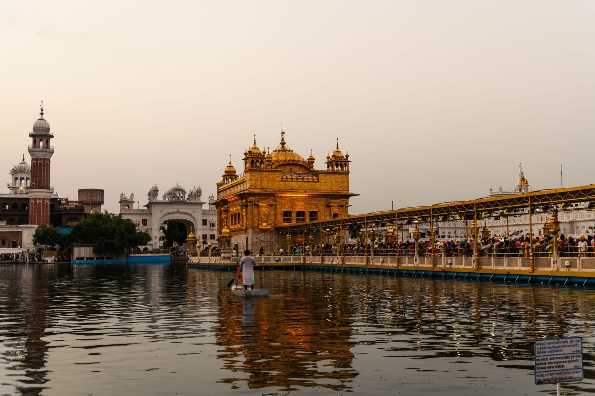 The Golden Temple at dusk, its gilded domes reflected in the still waters of the Amrit Sarovar, surrounded by white marble walkways and devotees in prayer