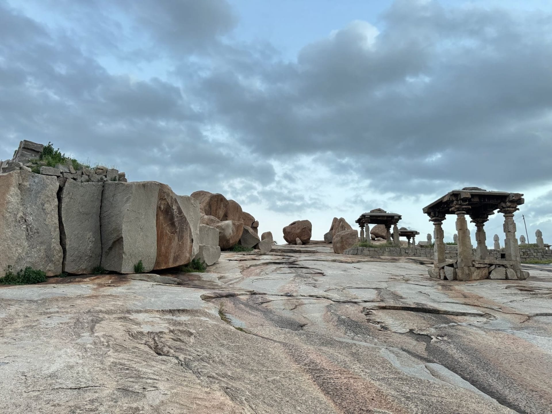 Ancient granite boulders stacked in impossibly balanced formations above the Tungabhadra River, with the carved gopuram of the Virupaksha Temple rising through warm haze in the background at golden hour