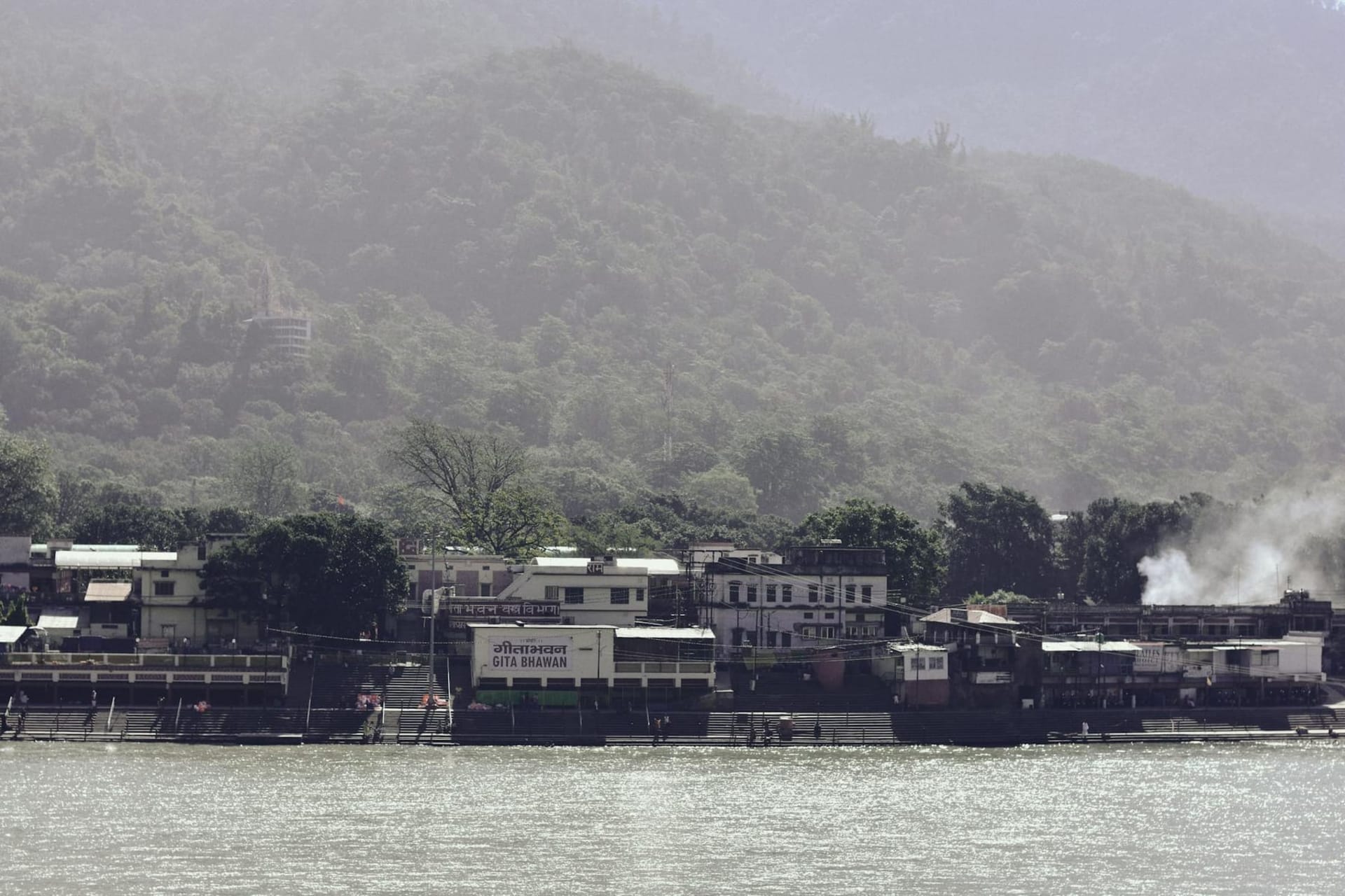 The Ram Jhula suspension bridge spanning the jade-green Ganges at Rishikesh, with ashrams and temple spires rising from the forested hillside behind it under hazy mountain light.