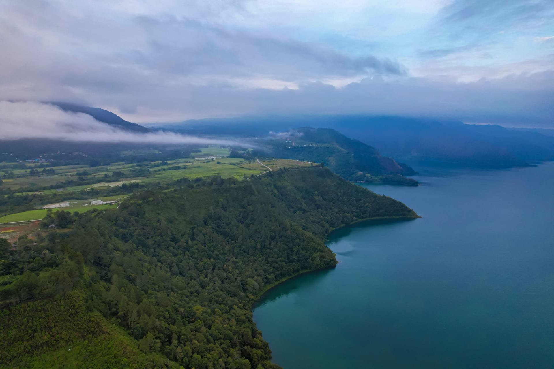 The huge expanse of Lake Toba seen from a hillside on Samosir Island, calm blue water ringed by green volcanic ridges under a wide sky