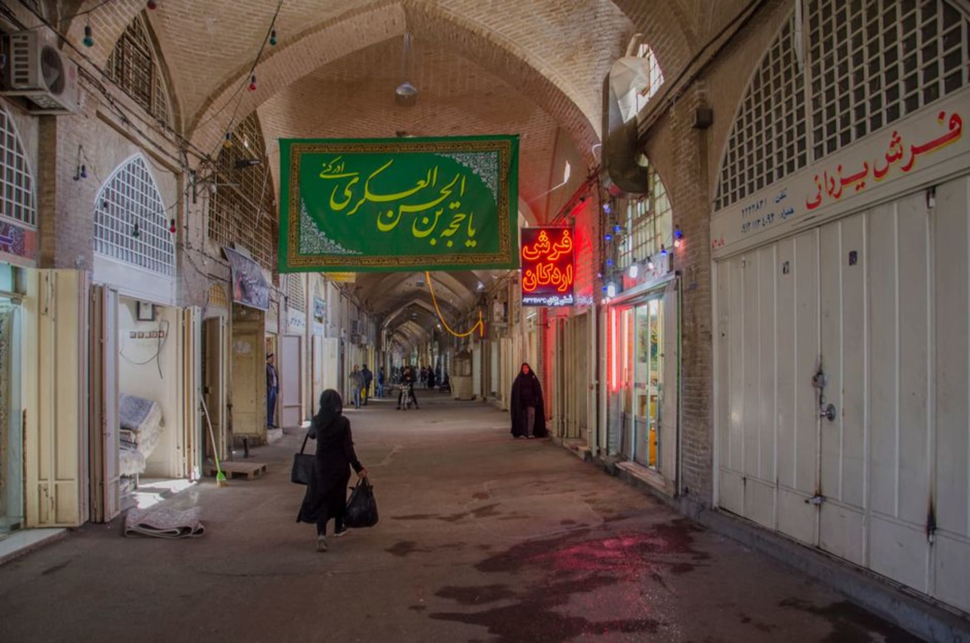 A vaulted brick corridor of the Isfahan Grand Bazaar lined with shop stalls and lit by shafts of light from ceiling openings