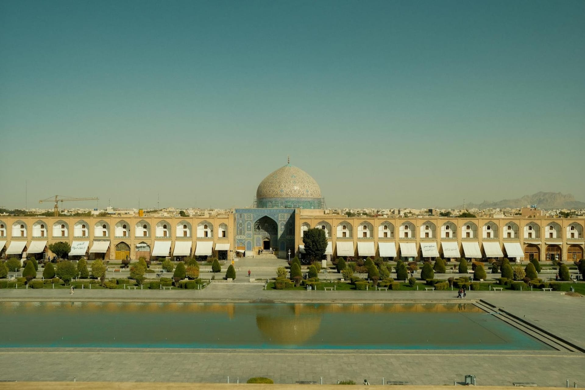 The vast expanse of Naqsh-e Jahan Square at dusk, the twin minarets of Masjed-e Shah reflected in the central pool, its turquoise faience glowing amber and cobalt in the fading light.