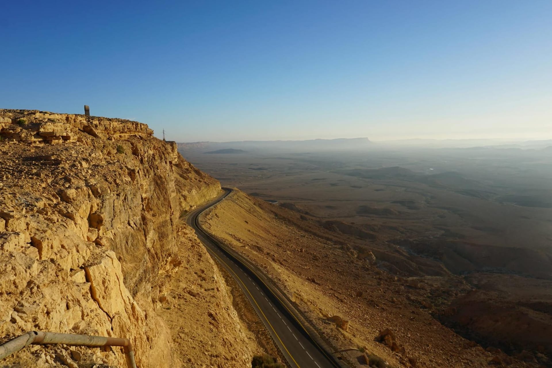 Aerial view of Makhtesh Ramon's vast ochre and rust-colored crater floor stretching to layered sandstone cliffs under a cloudless blue sky in the Negev Desert