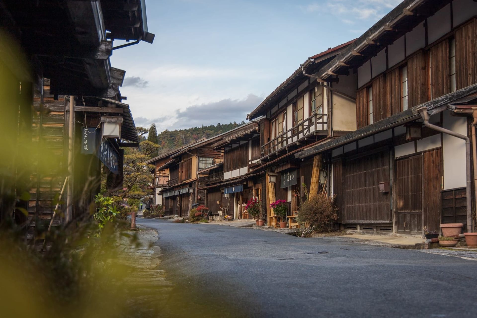 A narrow stone-paved street in Tsumago lined with dark-timbered wooden machiya houses, paper lanterns glowing amber under a grey mountain sky