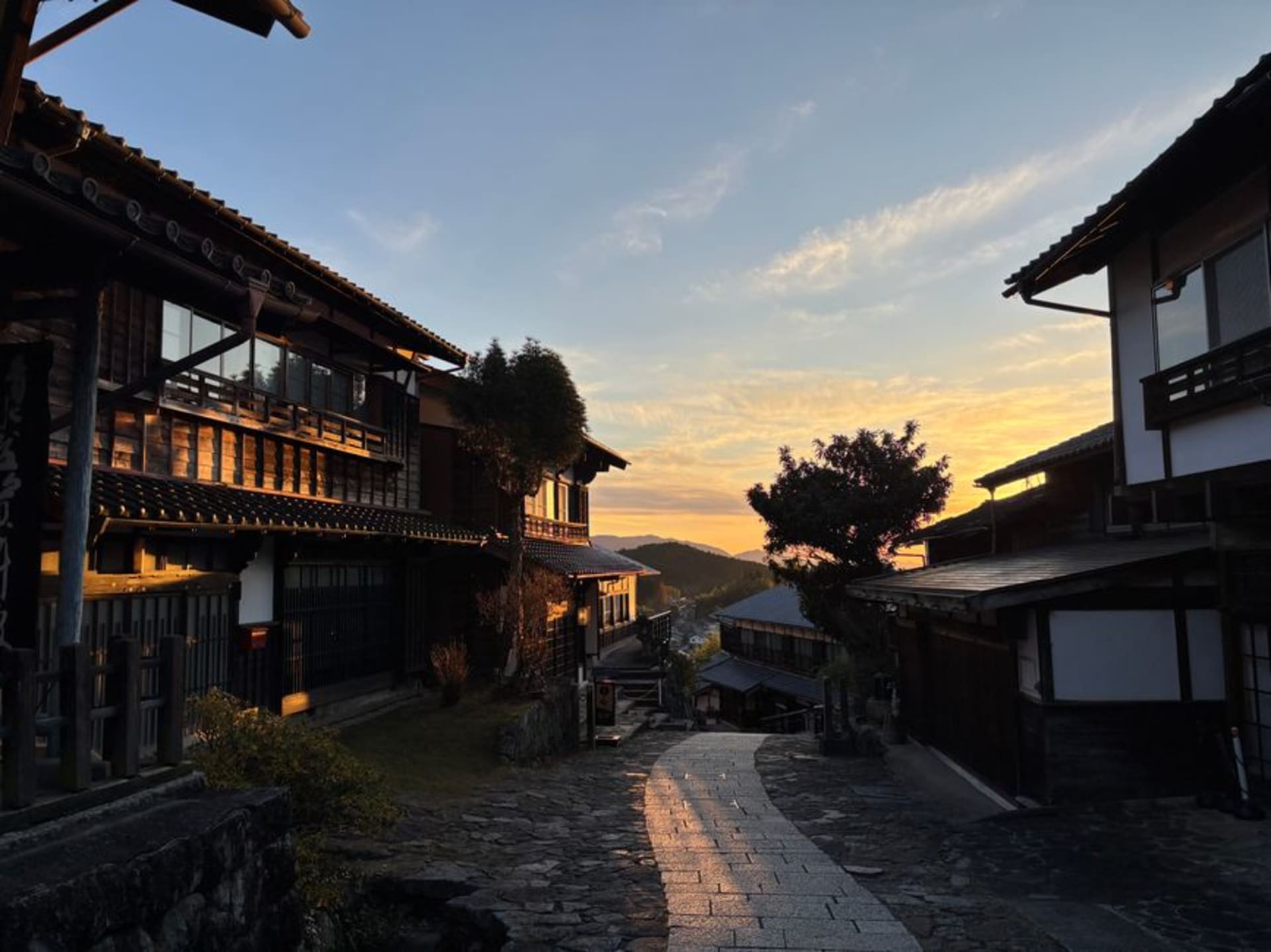 Magome's steep stone-paved main street rising between dark wooden inns and shops, a water wheel turning in the foreground under a mountain sky