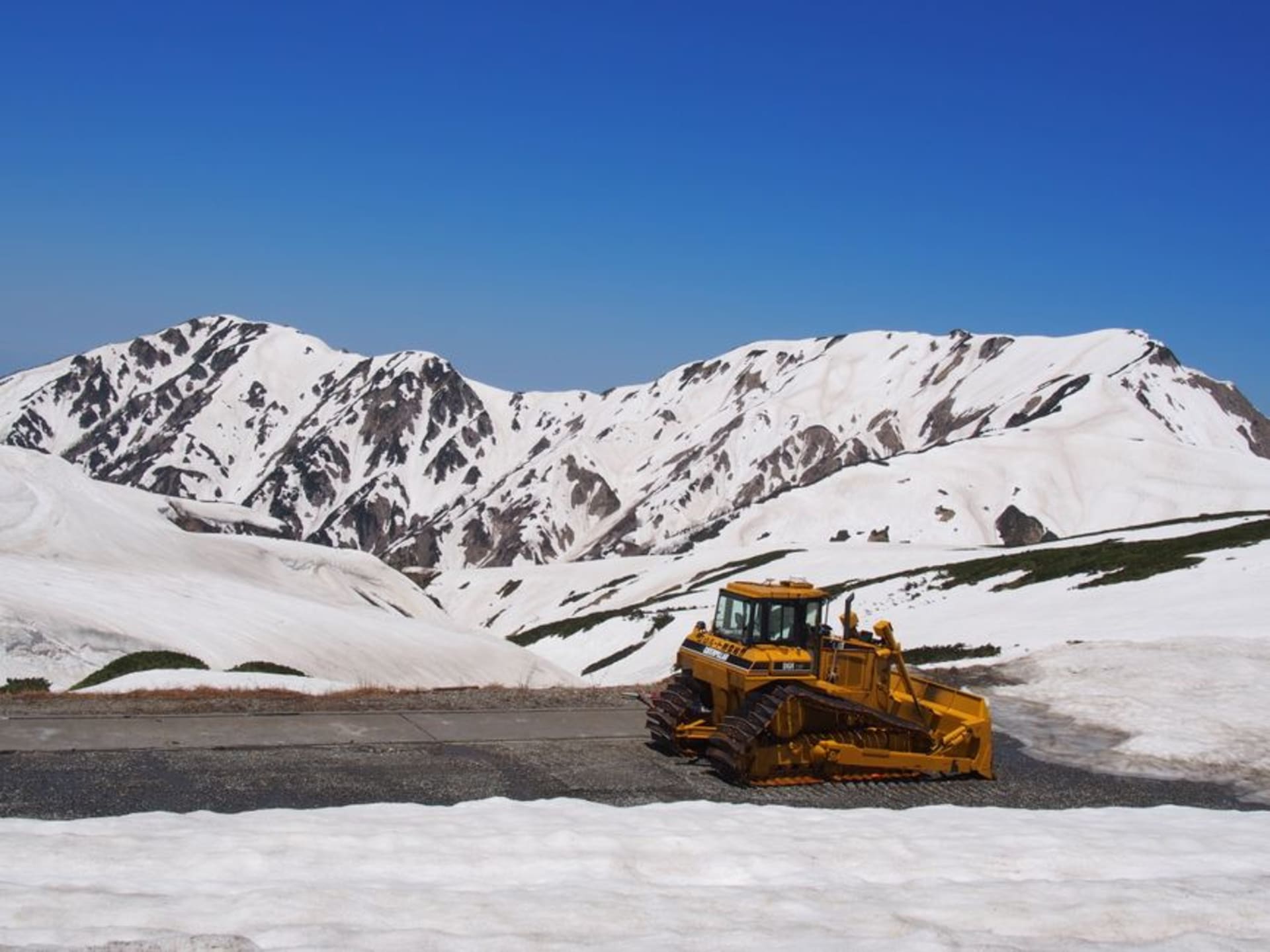 A bus threading between towering walls of snow far higher than the vehicle on the Tateyama Kurobe snow corridor under a blue sky