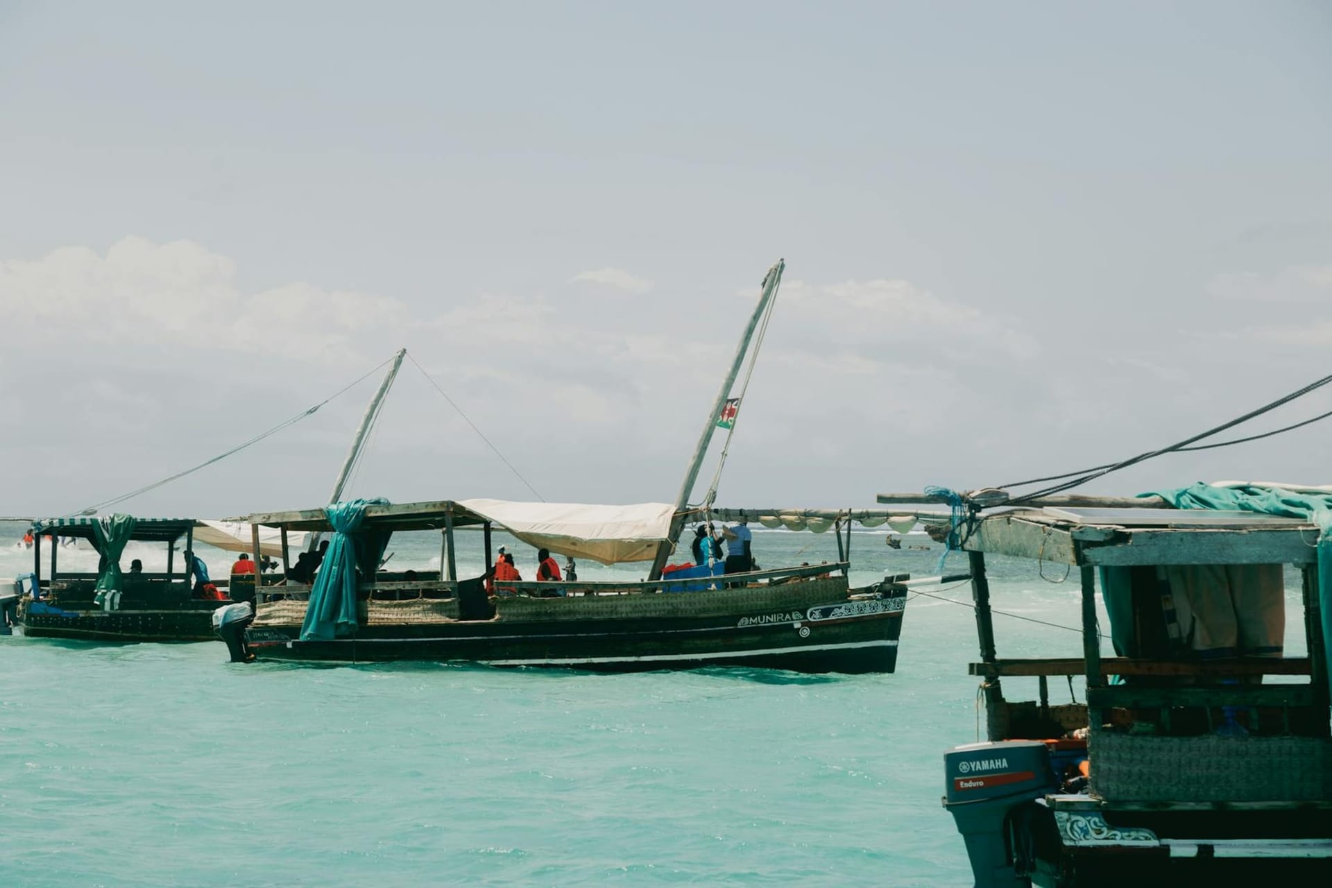 A weathered dhow sailing past the stone waterfront of Lamu Old Town at golden hour, mangrove poles stacked along the dock and whitewashed coral-stone buildings rising behind