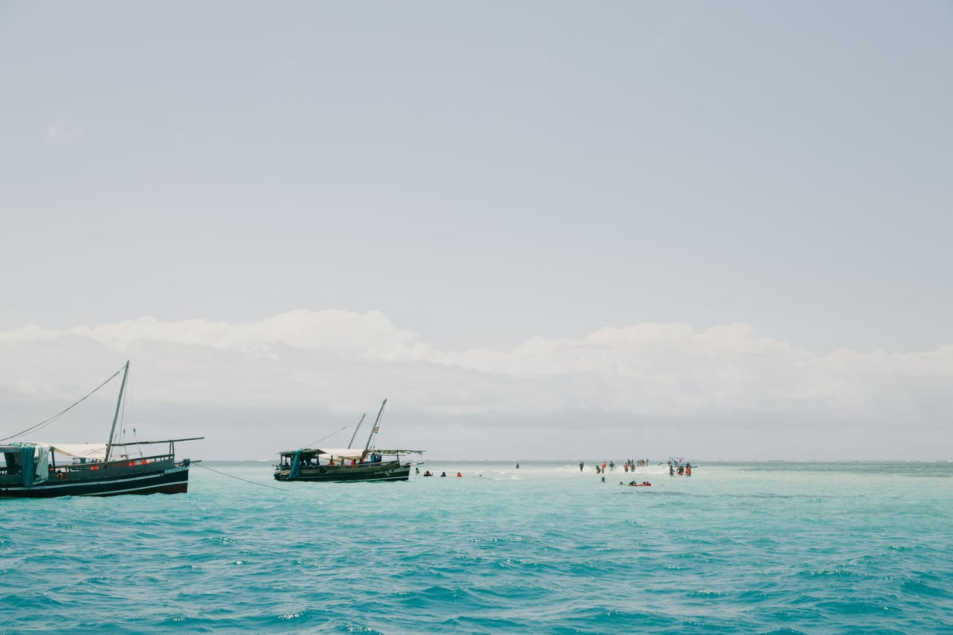 A wooden dhow with a rust-colored lateen sail gliding across the turquoise channel of Lamu harbor, with the whitewashed coral-stone buildings of the old town rising behind the waterfront palms.