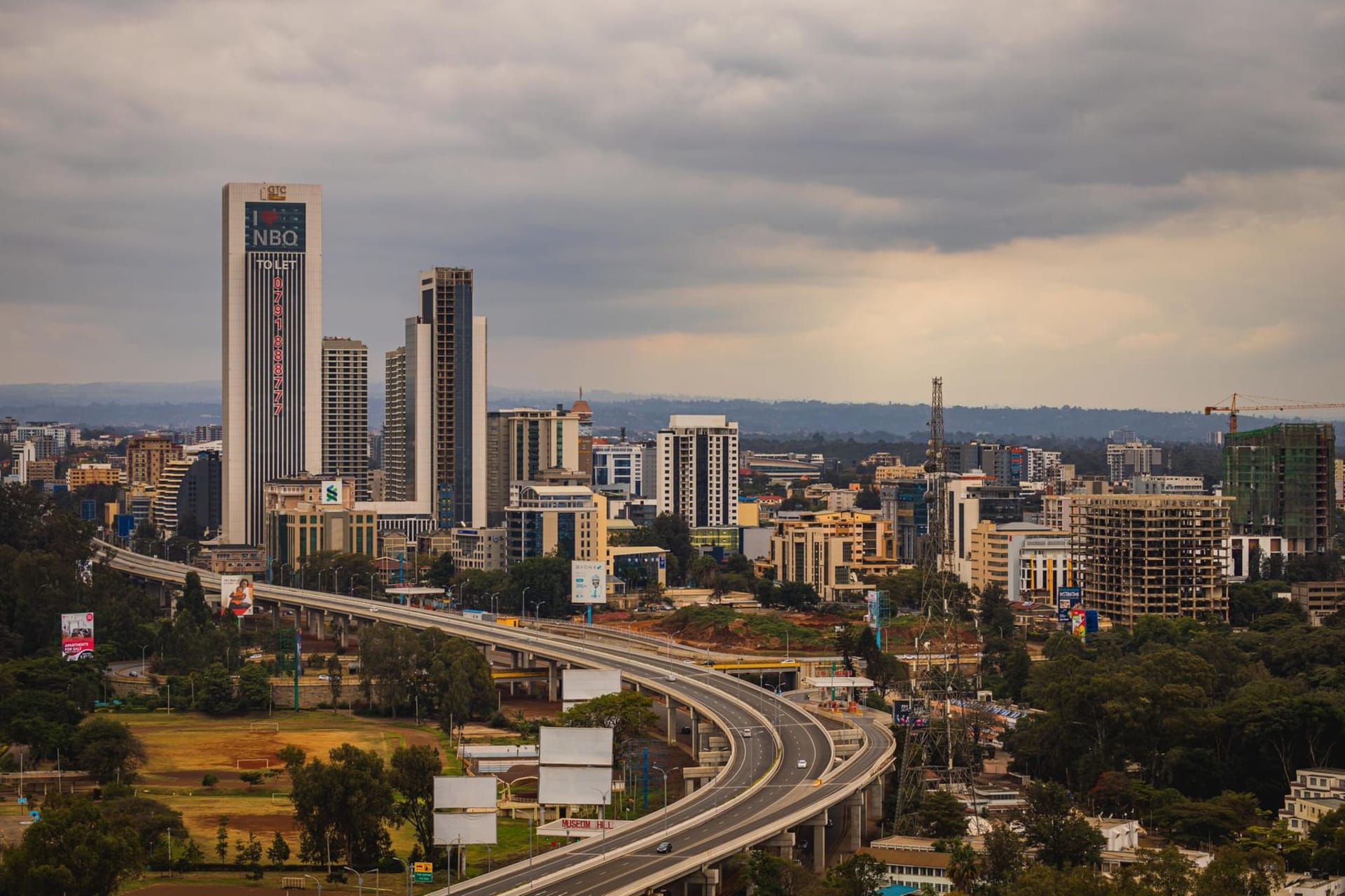 Nairobi's modern glass-tower skyline rising behind the golden savanna grasses of Nairobi National Park, a giraffe silhouetted against the city at dusk