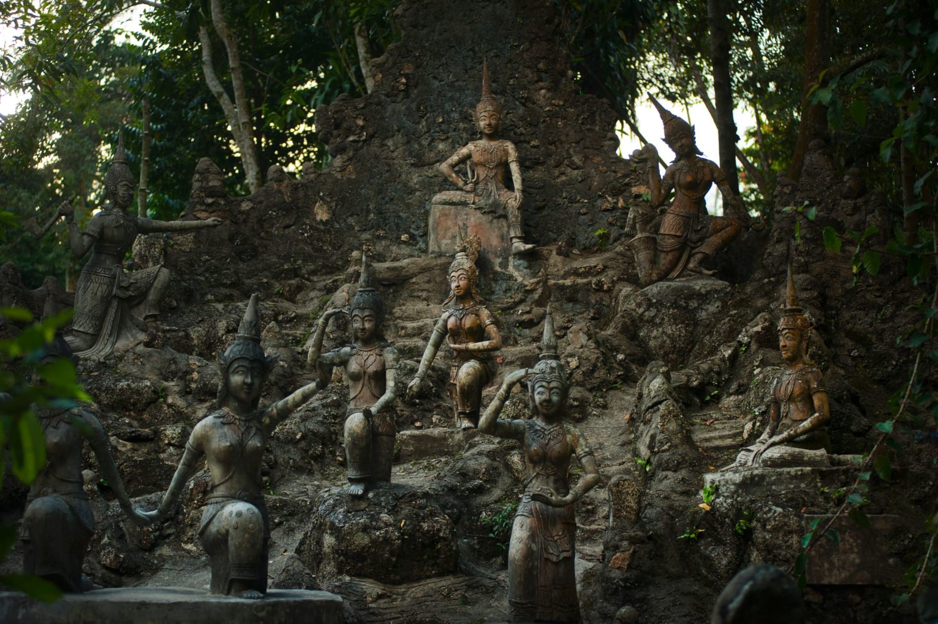 Moss-covered stone statues of deities and figures arranged among boulders, ferns and a small stream in the jungle of the Secret Buddha Garden on Koh Samui