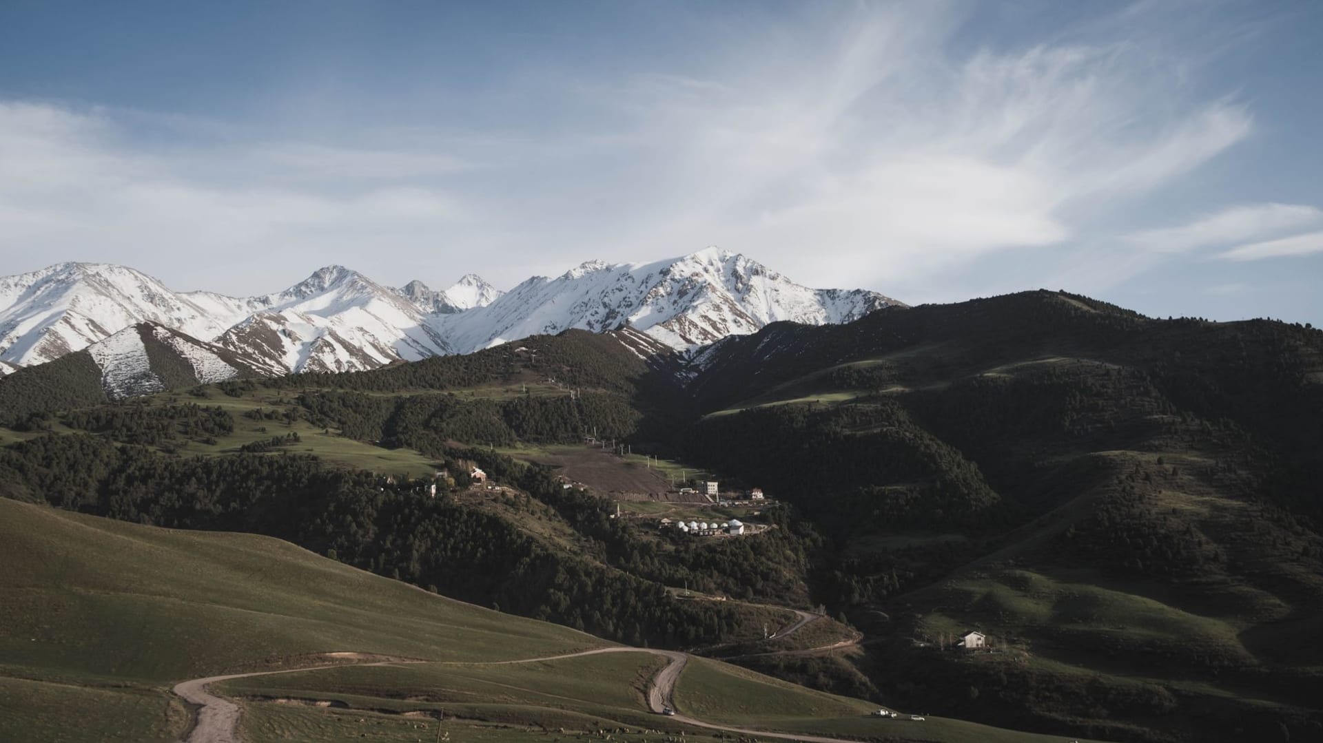 El amplio valle verde de Jyrgalan a pleno verano con un campamento de yurtas en el prado y picos escarpados atrapando la última luz