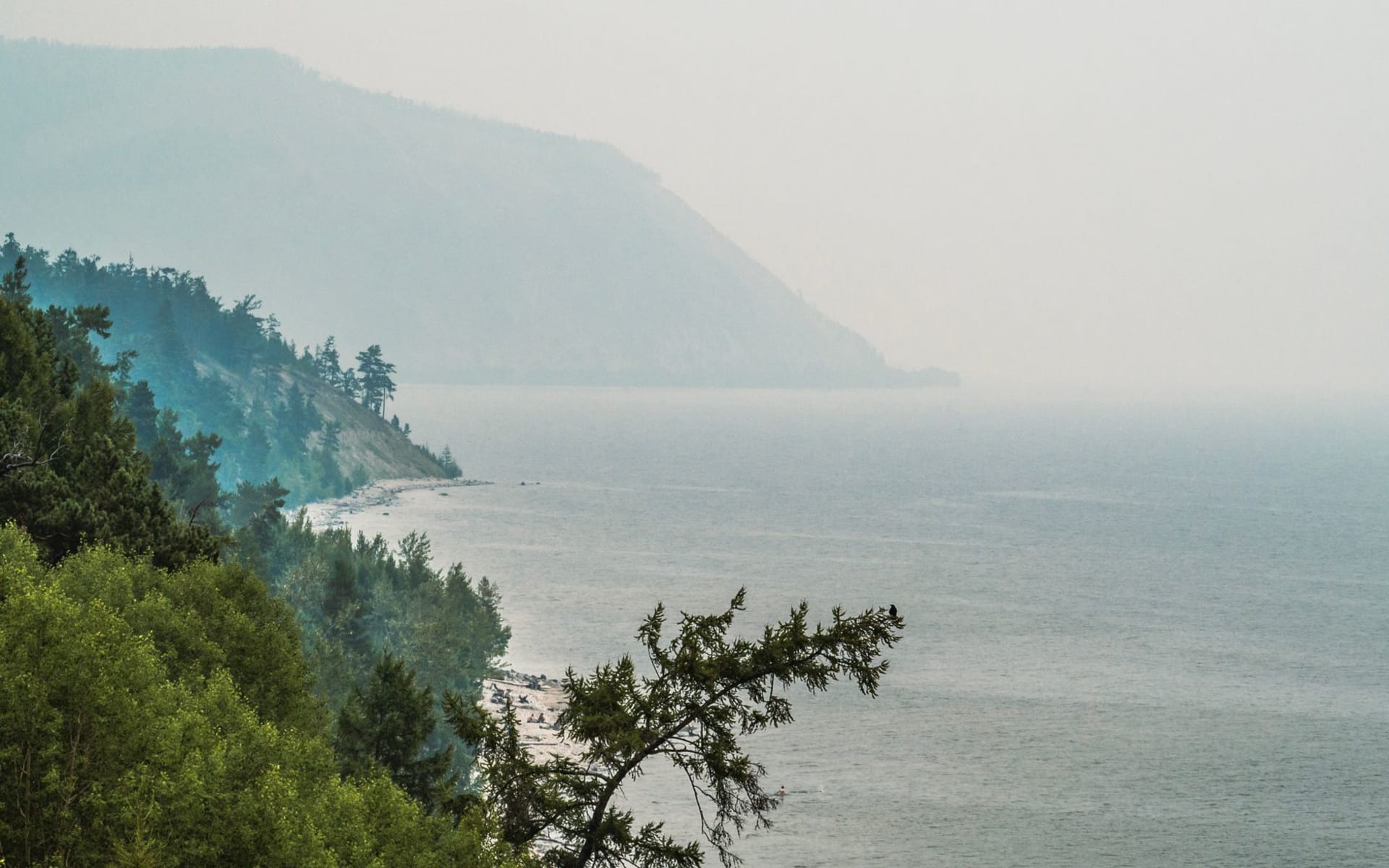 A vintage locomotive emerging from a tunnel cut into cliff rock directly above the glittering blue surface of Lake Baikal