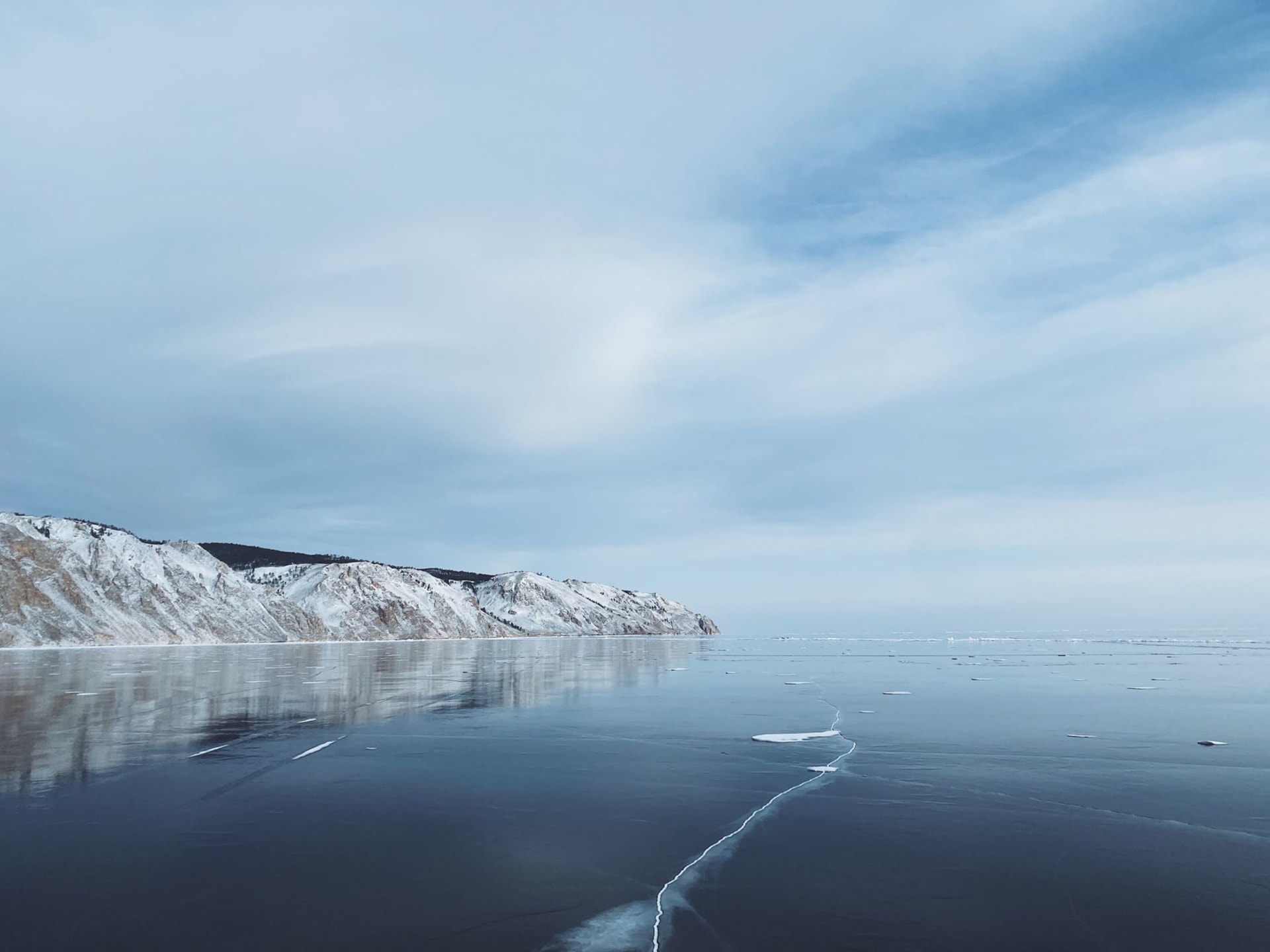 The vast frozen surface of Lake Baikal under an expansive blue sky with snow-dusted mountains in the distance