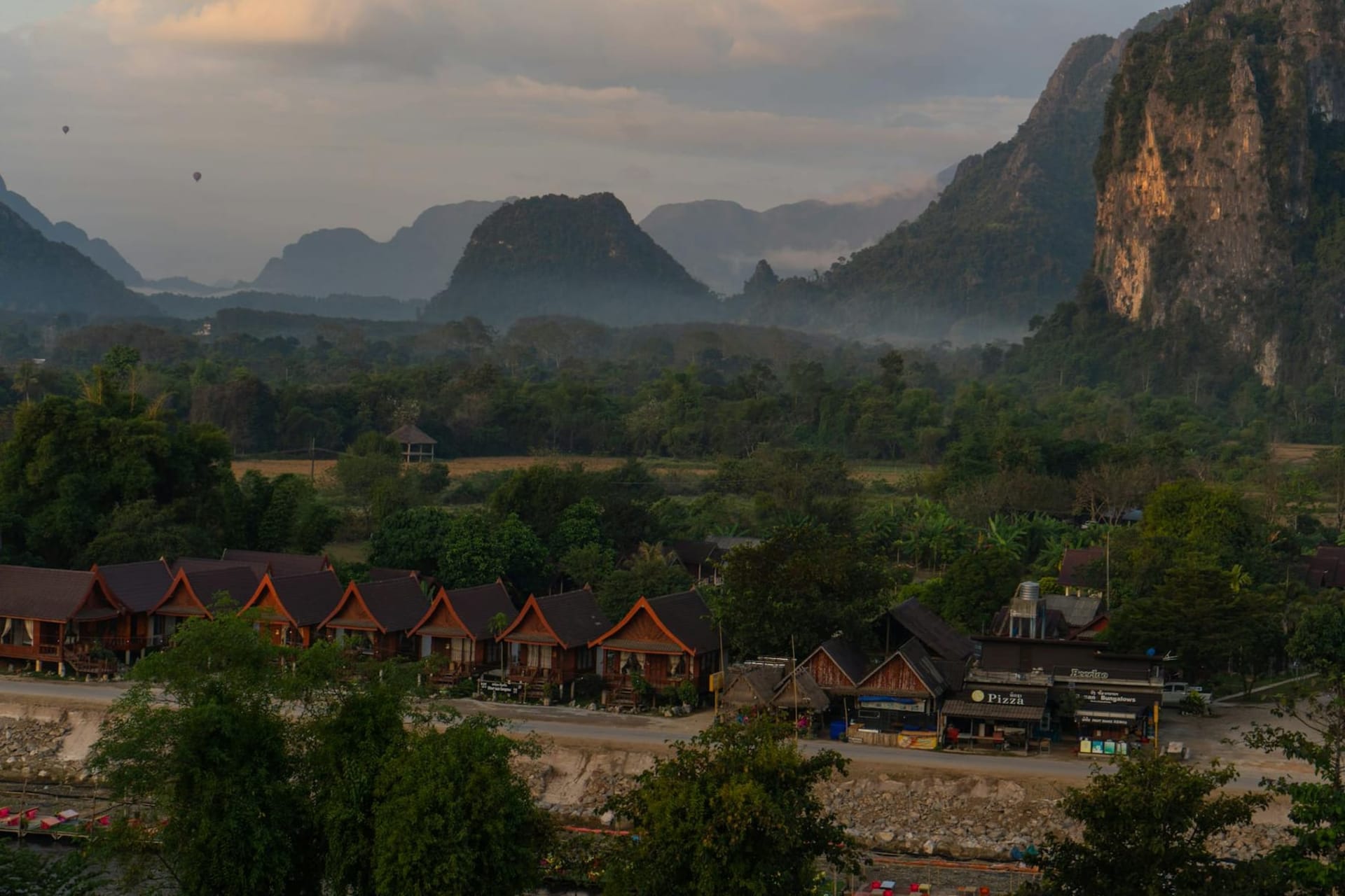 Dramatic limestone karst peaks rising above the Nam Song River at golden hour, with a colorful hot-air balloon drifting between the cliffs and a wooden longtail boat moving slowly along the glassy water below.