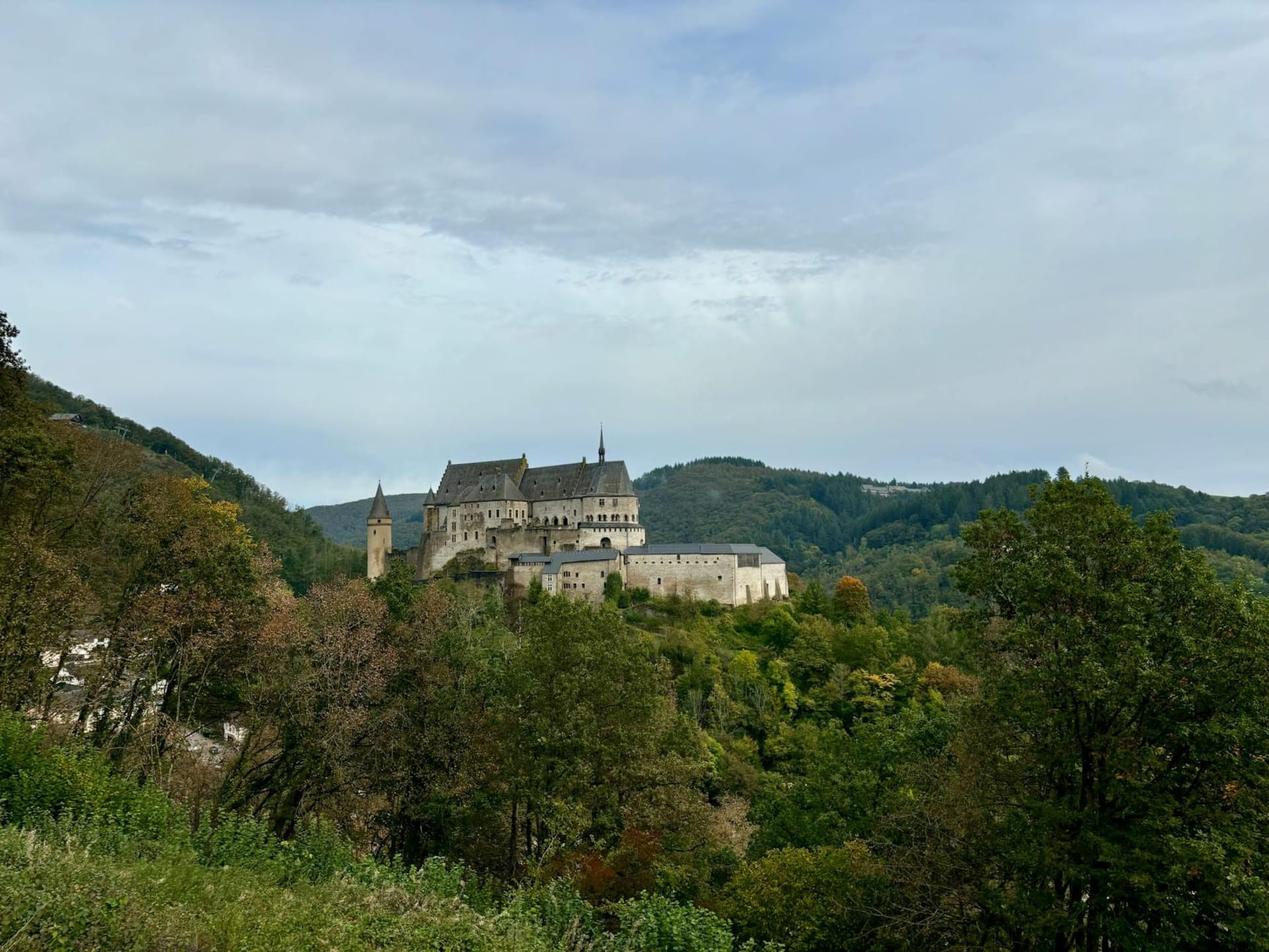 Vianden castle rising above a dense cluster of slate-roofed houses in the Our river valley, framed by wooded hillsides under a pale morning sky