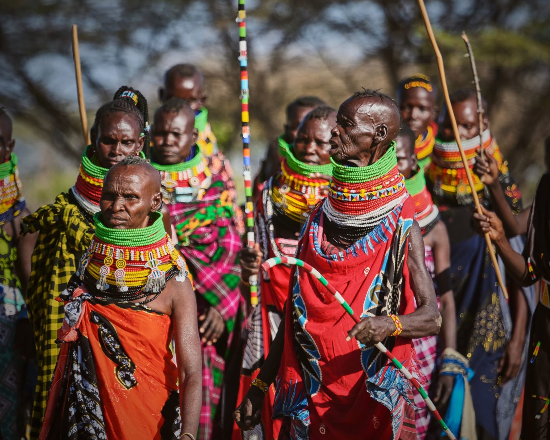 A dusty main street in Talek with Maasai women in red shukas passing a row of small shops and safari vehicles