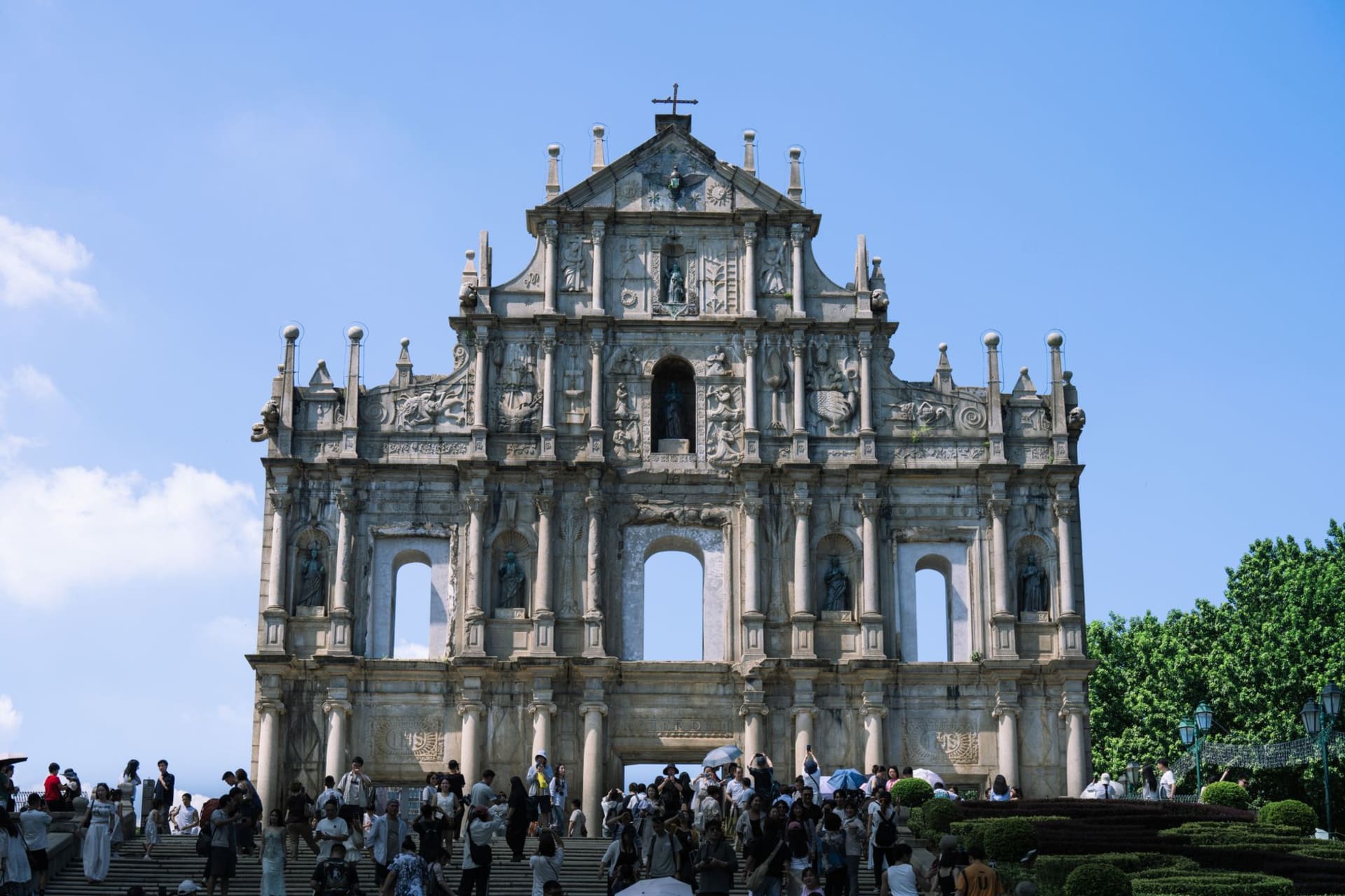 The roofless stone facade of the Ruins of St. Paul's rising against a clear blue sky, with visitors gathered on the steps below