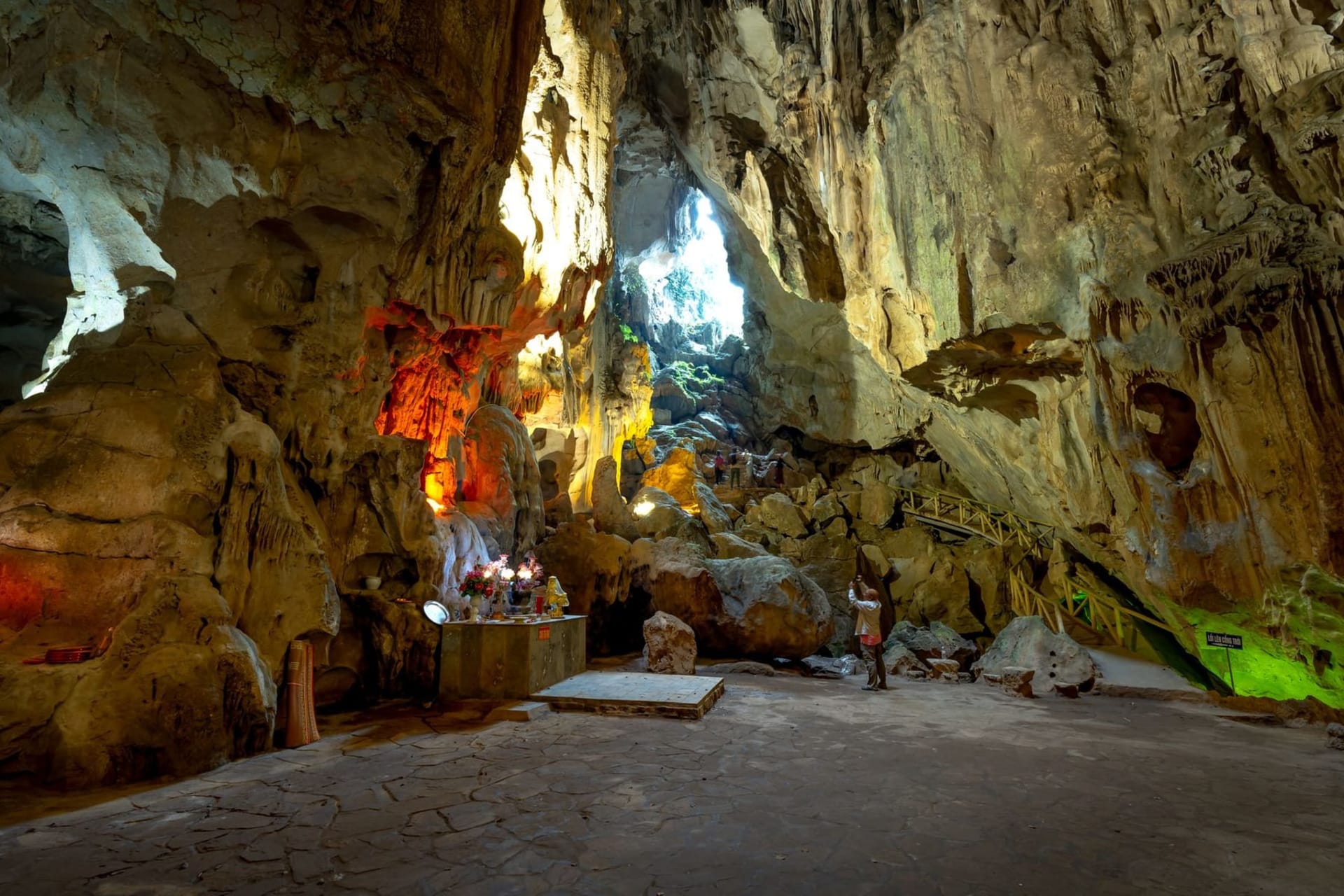 A vast limestone cave entrance at Gunung Mulu National Park, with a wooden boardwalk leading into darkness and shafts of pale green light filtering through jungle canopy overhead.