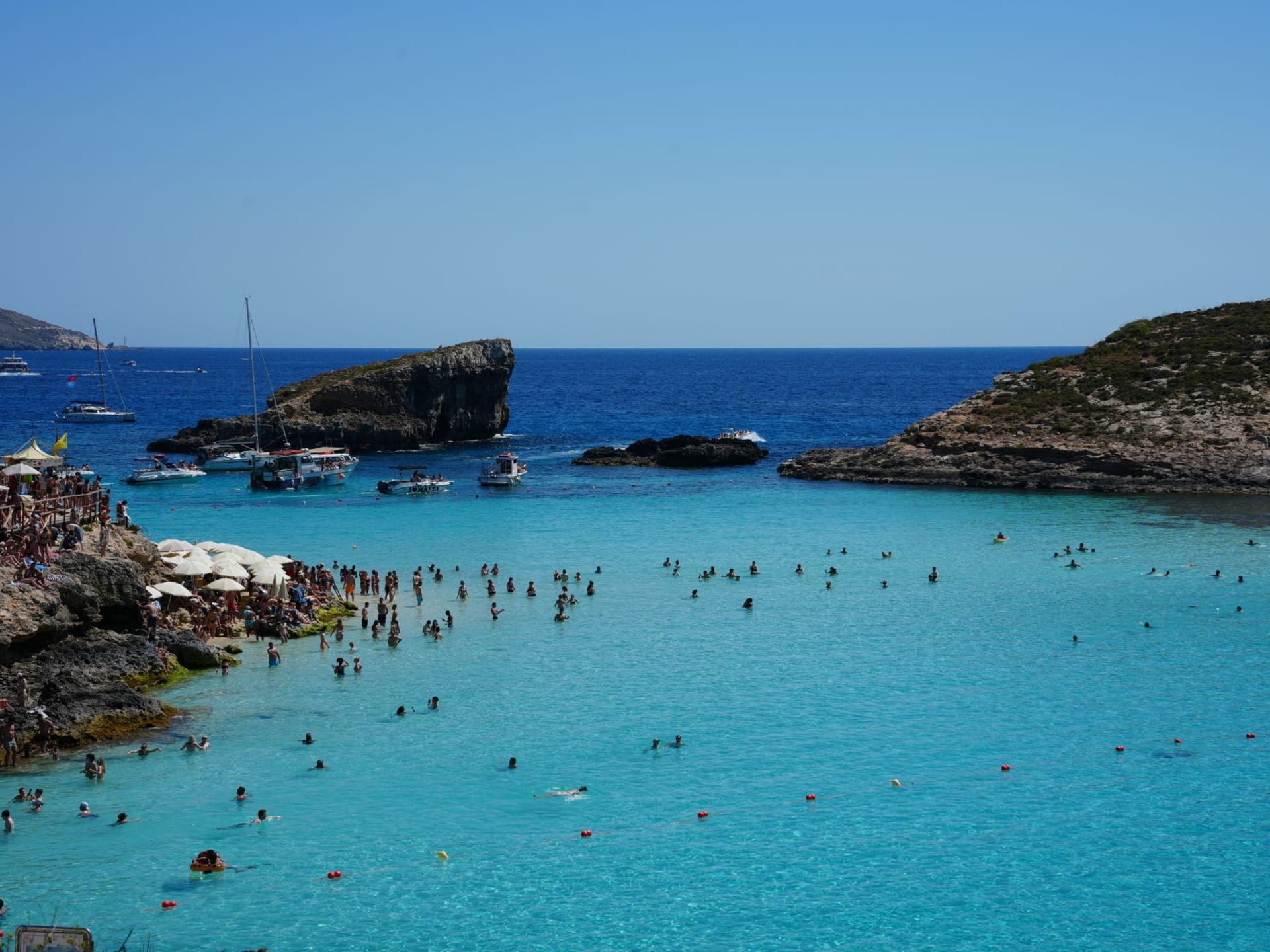 Comino's Blue Lagoon with crystal-clear turquoise water, white limestone seabed visible through the shallows, and a few small boats anchored in the bay
