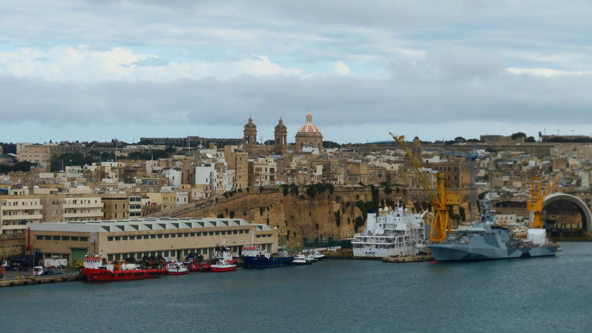 El horizonte del frente marítimo de Senglea visto desde el Gran Puerto, con barcas de pesca y luzzu tradicionales amarrados junto a las murallas fortificadas de color miel, Malta.