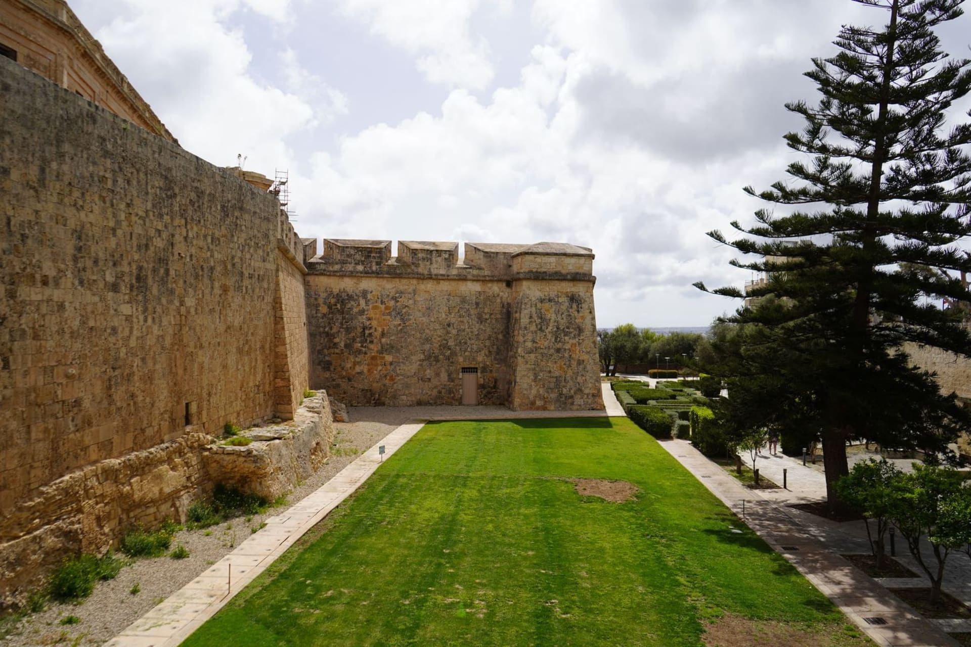 Narrow sun-bleached limestone lane cutting between high medieval walls in Mdina, Malta, with ornate baroque doorways and a bell tower rising against a pale blue sky