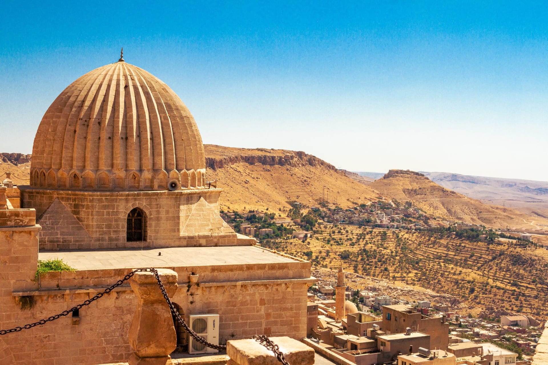 The grand temple complex of Hatra, massive stone iwans and Corinthian columns rising from the desert floor, Parthian arched halls blending with classical colonnades under a blue sky