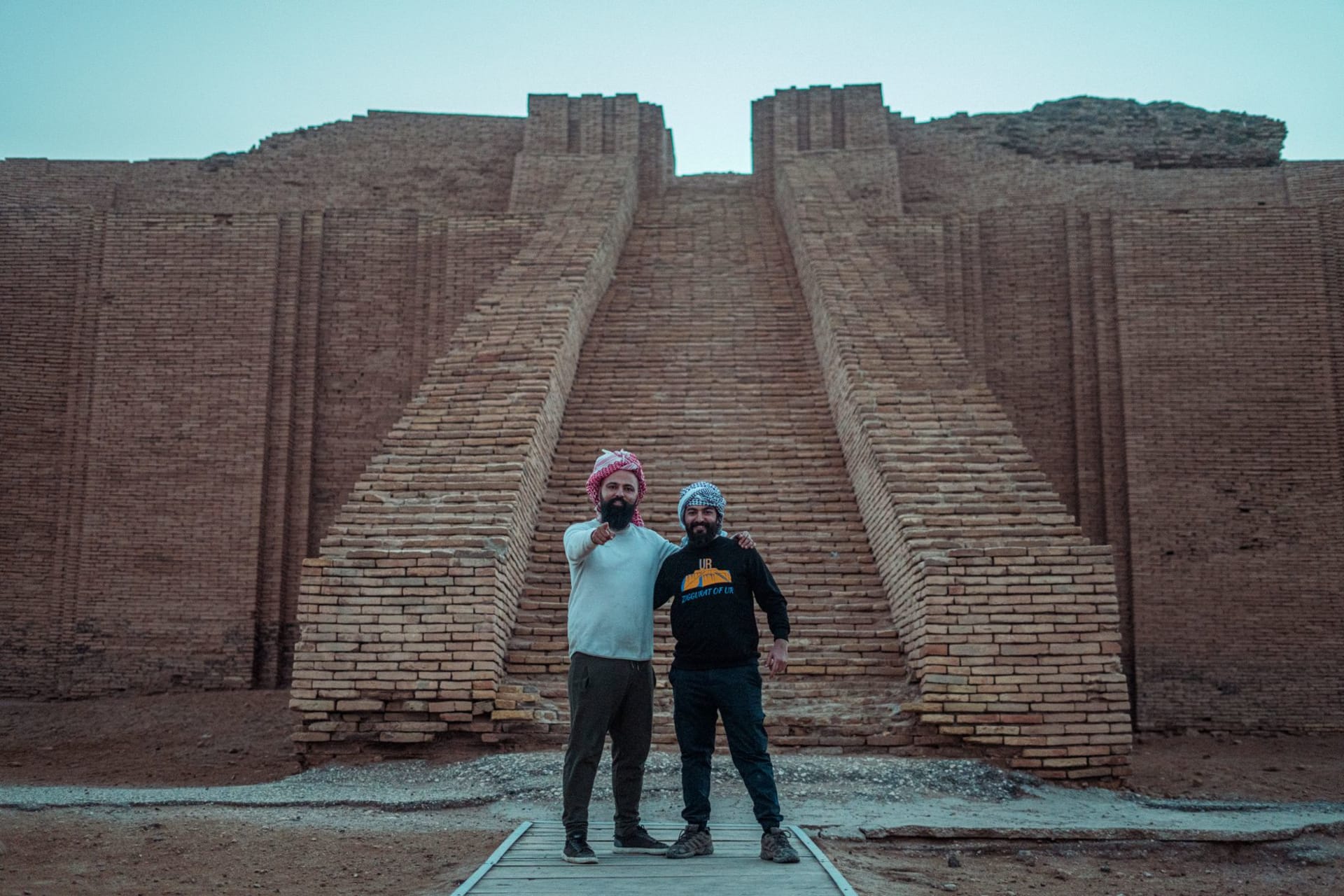 Two tourists standing at the base of the ancient Ziggurat of Ur, a massive stepped pyramid of sun-baked brick rising from the flat desert plain of southern Iraq