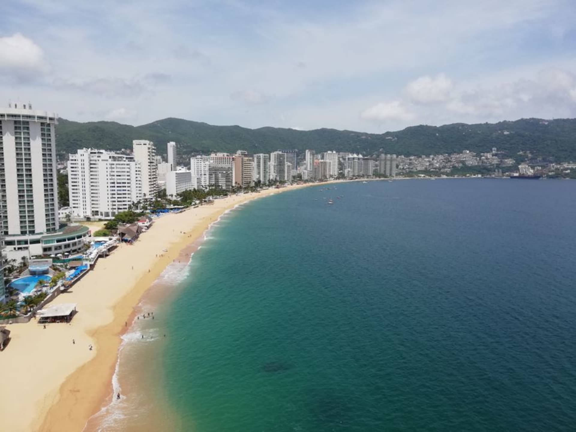 Acapulco's horseshoe bay at sunset, the Sierra Madre mountains behind the city, the curve of hotels along the Costera visible, and the Pacific Ocean turning gold in the fading light