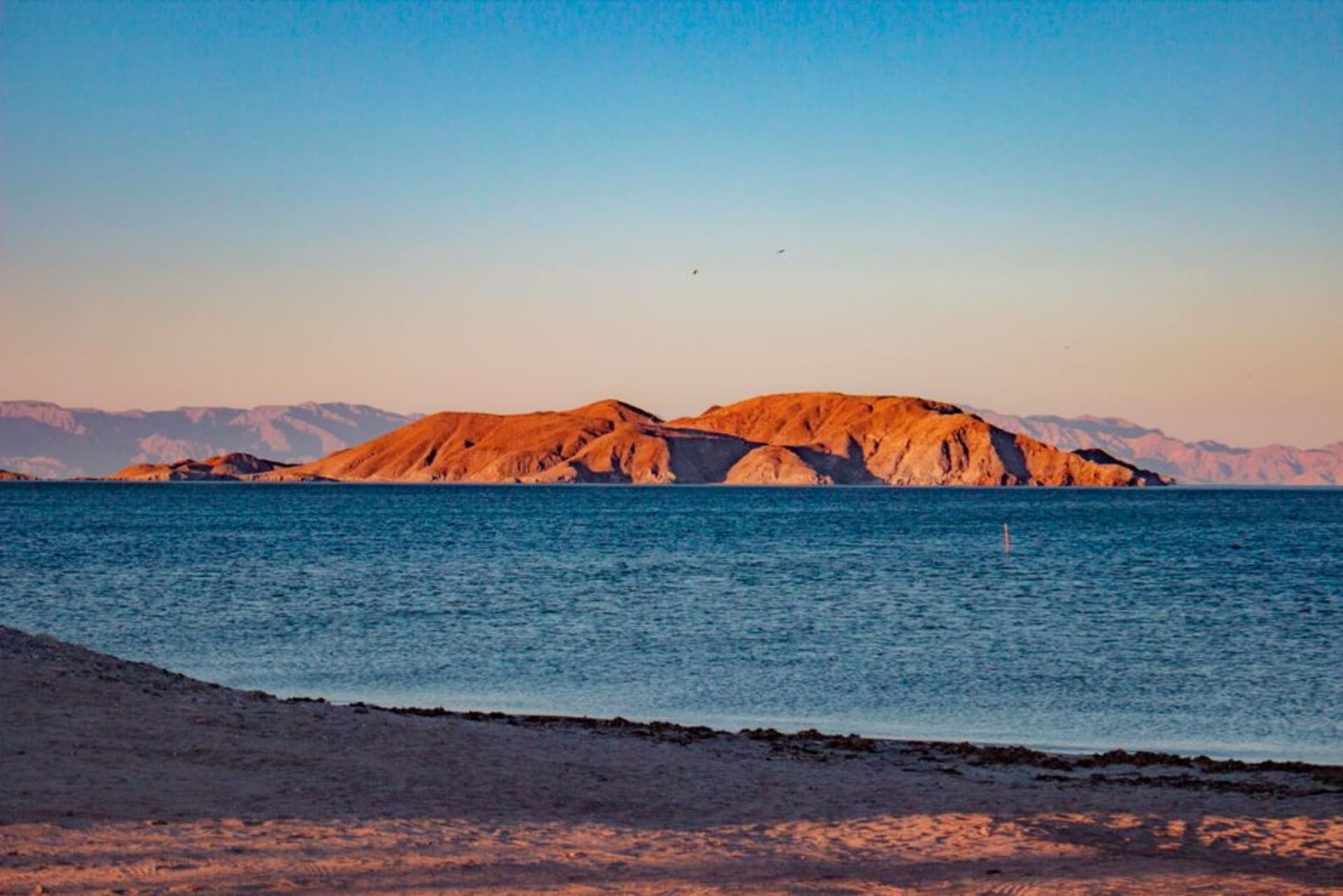 A wide turquoise bay ringed by volcanic hills at golden hour, with dark island silhouettes stacked across the water toward the horizon