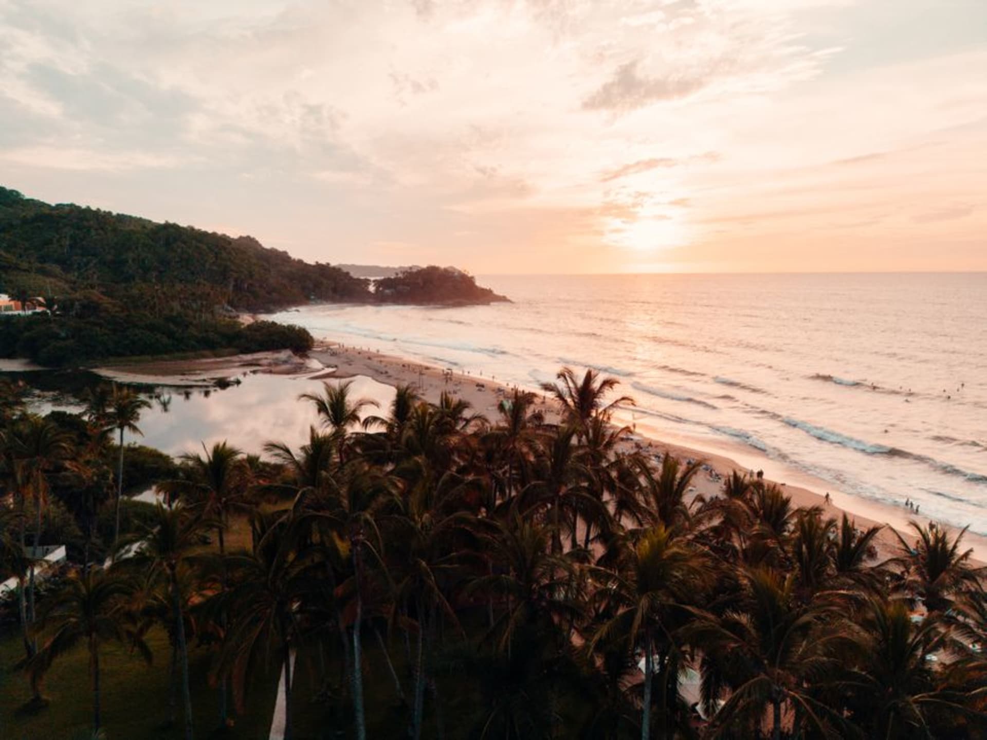 Sweeping arc of Bahía de Matanchén with surf breaking along the shore and dense jungle hills rising behind the beach under afternoon light