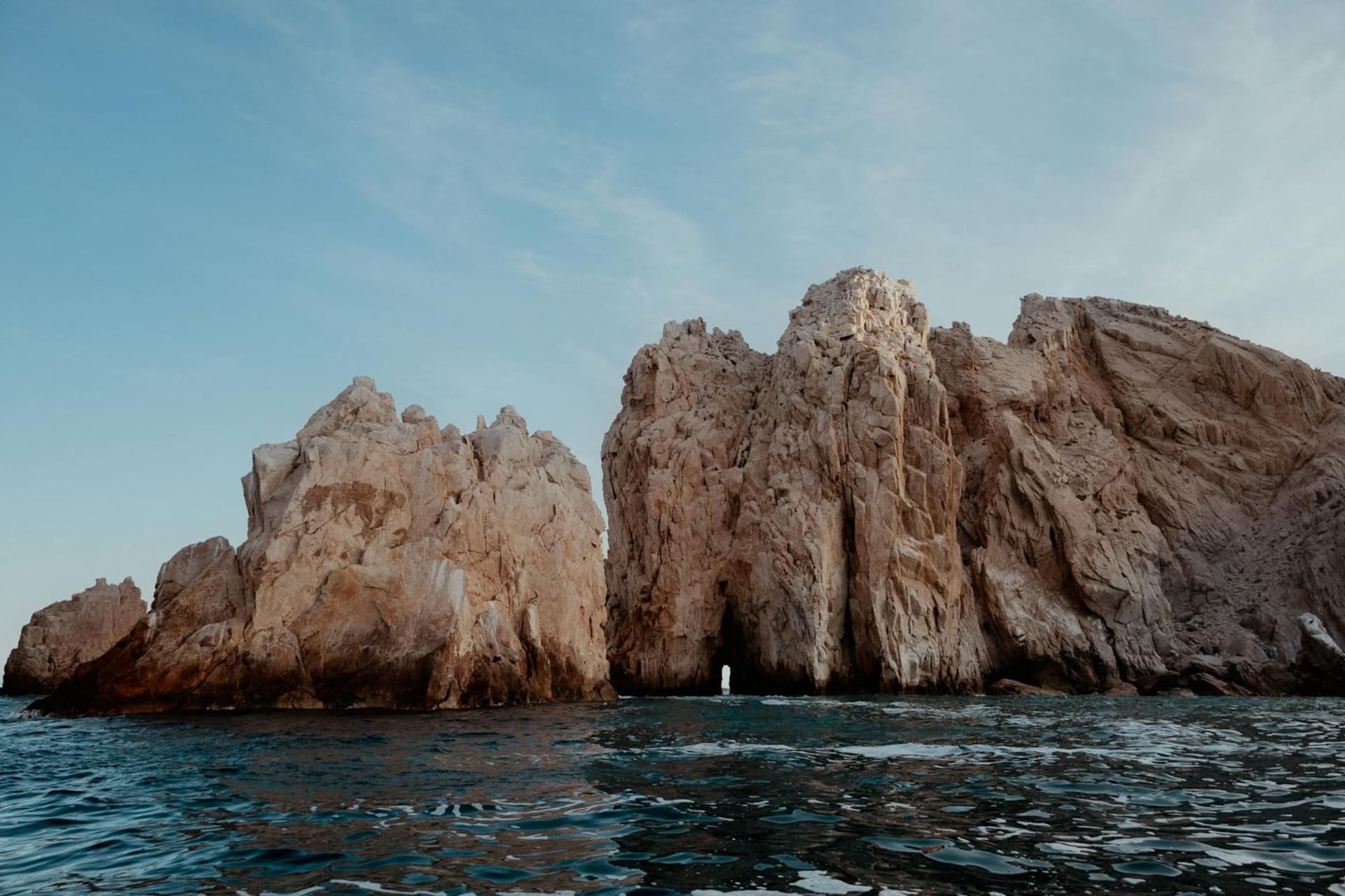 The iconic El Arco rock formation at Land's End in Cabo San Lucas, where the deep blue Pacific Ocean meets the turquoise Sea of Cortez, with pelicans resting on sun-bleached granite and a scattering of sea lions on the pale sand below.