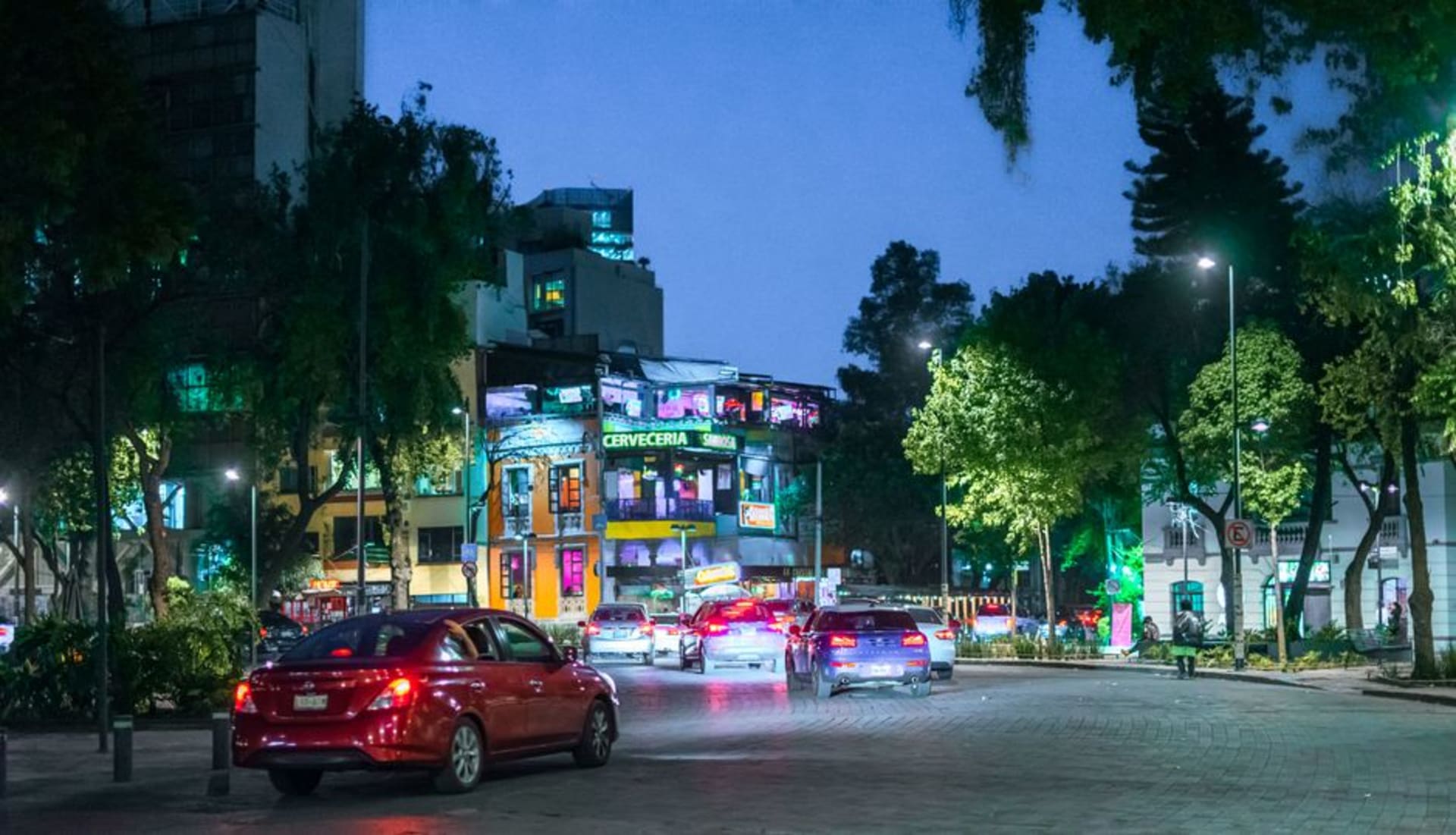 A tree-lined street in Colonia Juárez at dusk, softly lit restaurant terraces open to the sidewalk and pedestrians moving unhurriedly along Londres
