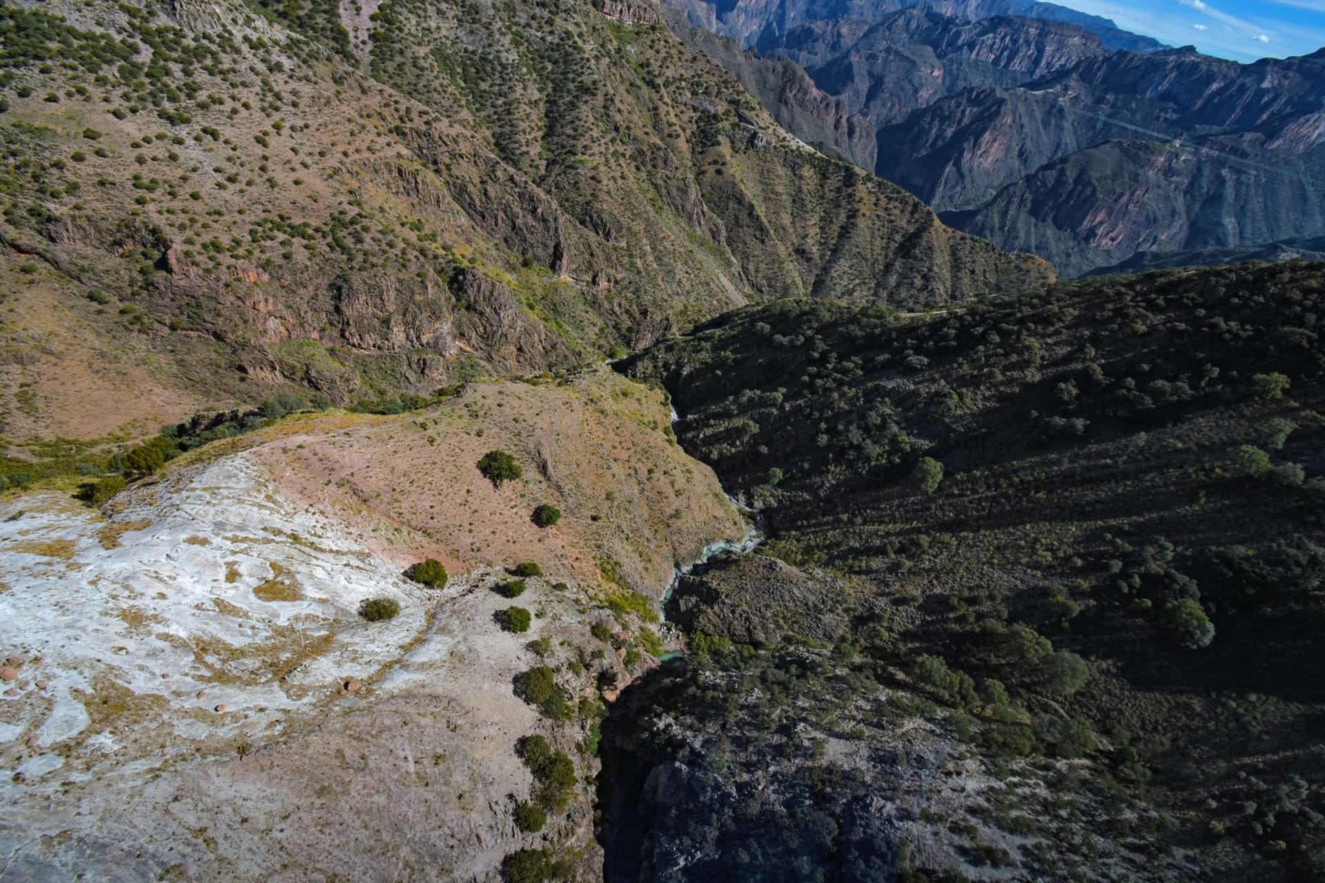 Aerial view of the vast Copper Canyon barrancas in Chihuahua, Mexico — layers of rust-red and pine-green canyon walls dropping into mist-filled gorges under a wide blue sky