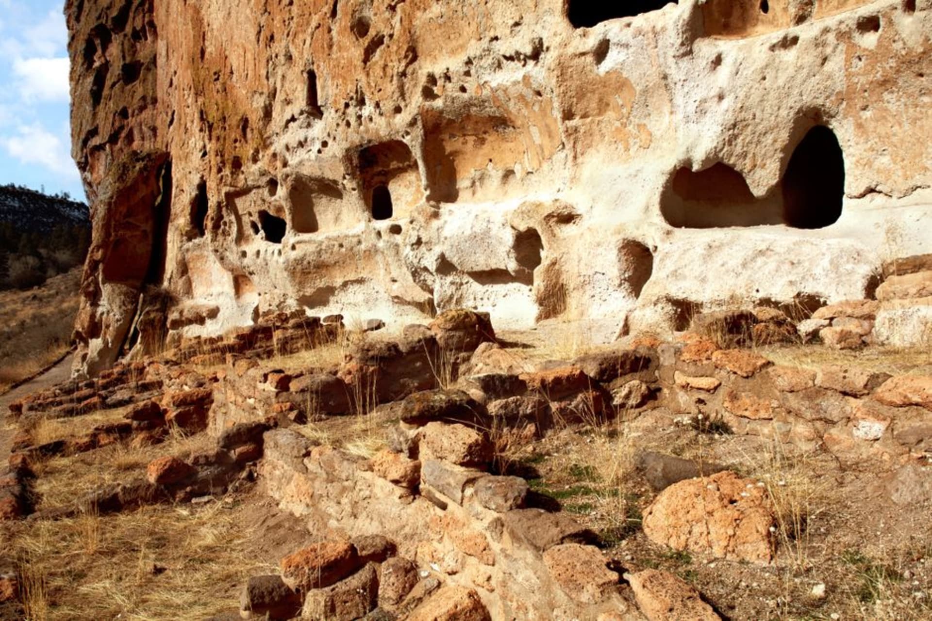 Multi-room stone structures tucked into a natural alcove high in the ochre cliffs of Cañón de la Huapoca, Chihuahua, against an open sierra sky