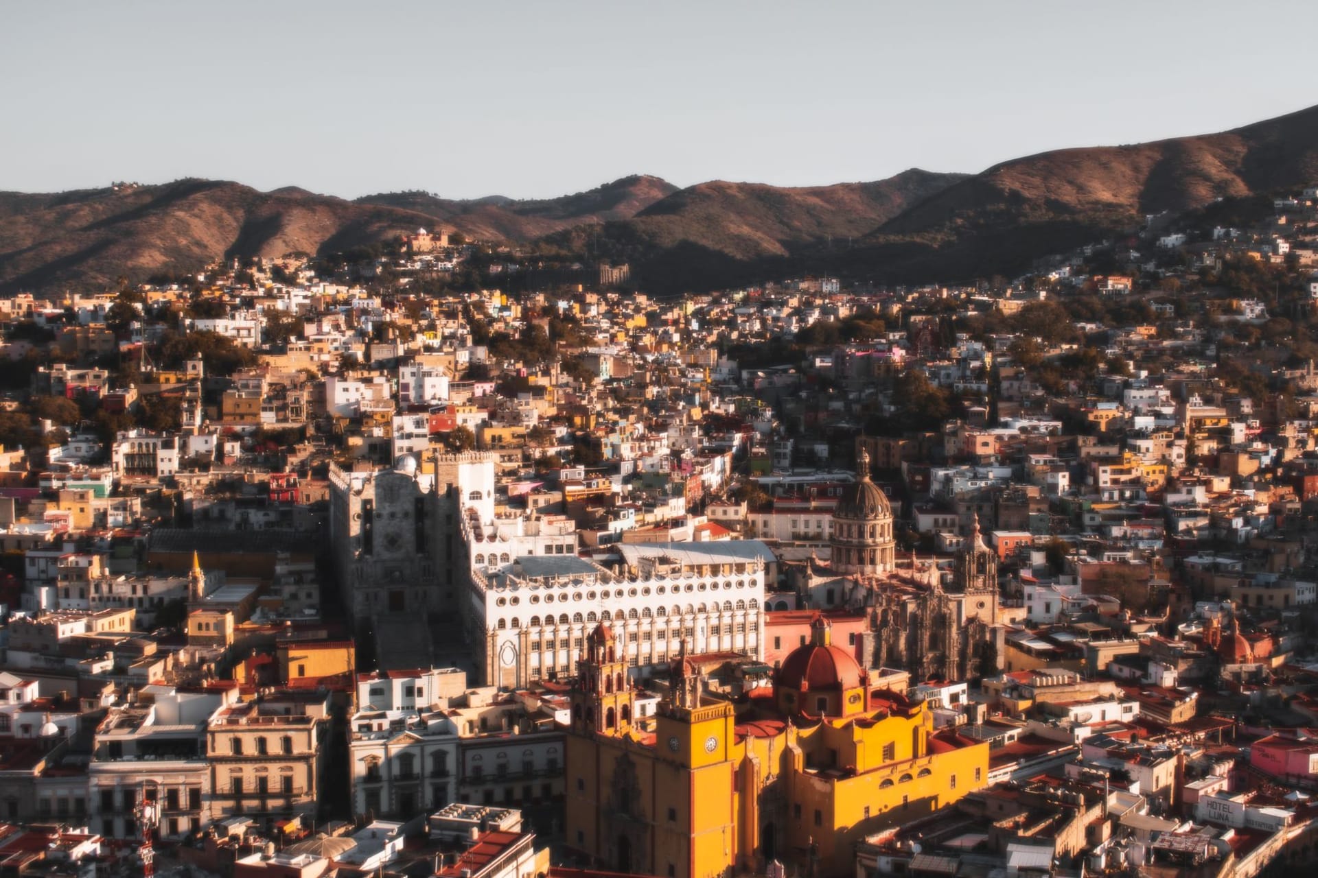 The hillside panorama of Guanajuato city, its buildings in layers of yellow, ochre, pink, and terracotta climbing the ravine walls, the Basílica visible at the center, mountains behind