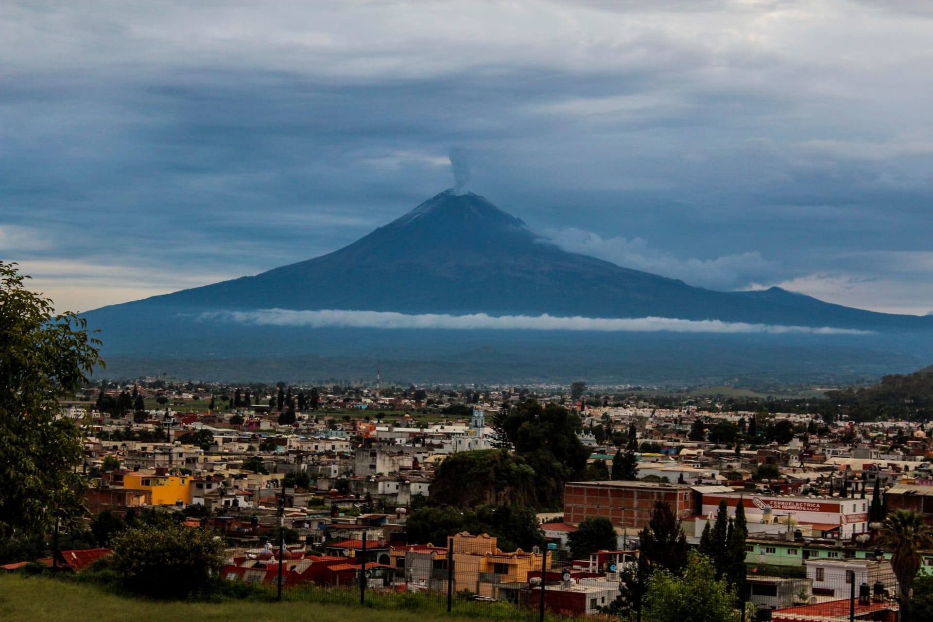 The stone church tower of San Juan Parangaricutiro rising from a frozen black lava field, the cone of Paricutín volcano visible in the distance behind it under a pale morning sky