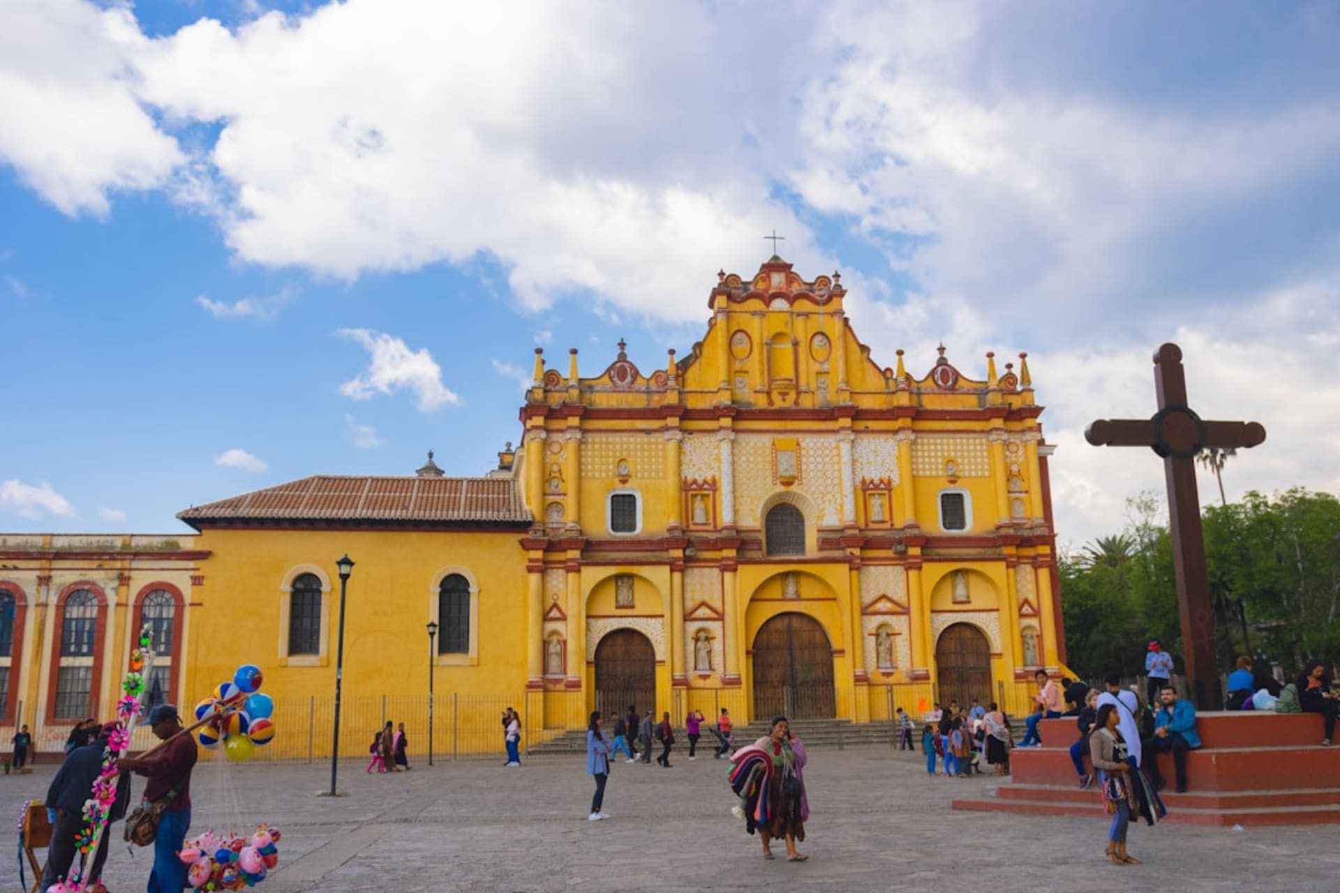 The white facade of the Church of San Juan Bautista rising above a pine-shaded plaza in San Juan Chamula, forested highlands visible in the distance