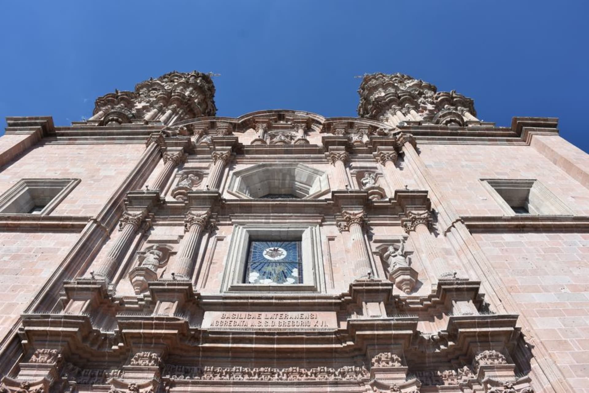The twin baroque towers of the Basílica de Nuestra Señora de San Juan de los Lagos rising above the plaza at dusk, their stone facades lit warm against a deep blue sky