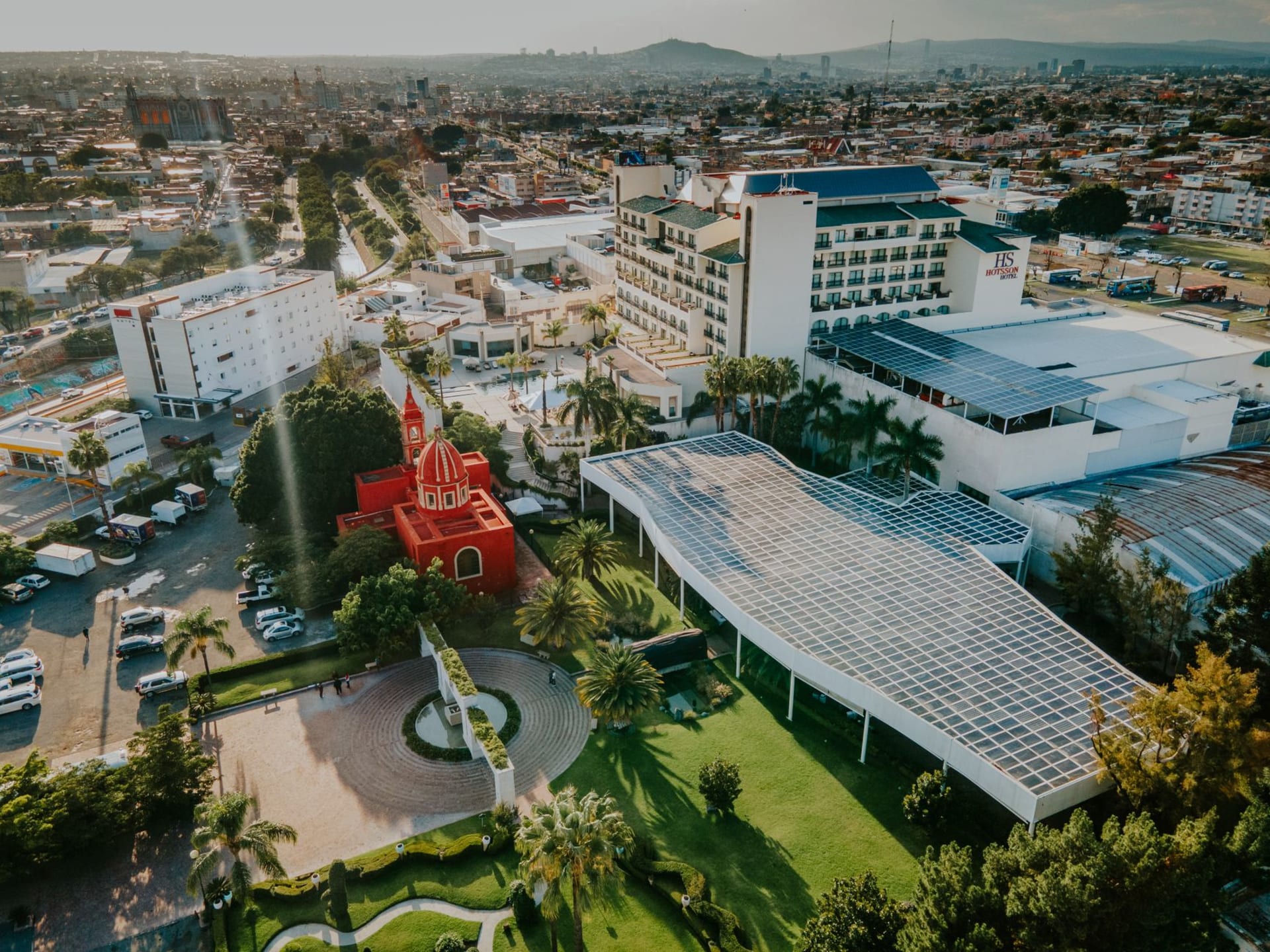 The main plaza of Tecate with the brewery building visible beyond the trees, a quiet afternoon with locals on benches and the border hills in the background
