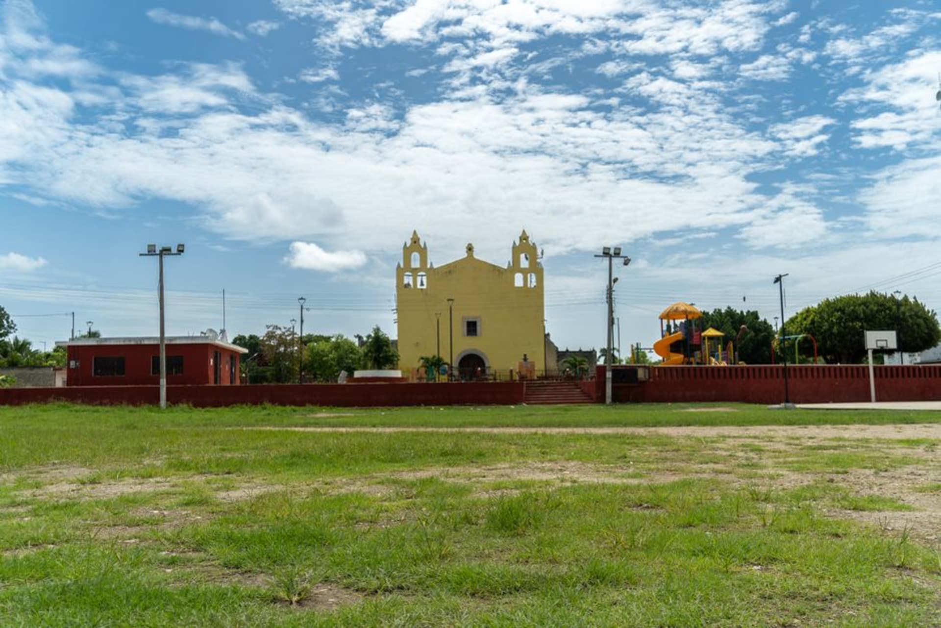 The wide plaza of Tekax at midday, the colonial facade of the Parroquia de San Juan Bautista rising above empty benches and a few palms casting narrow shadows on the stone paving